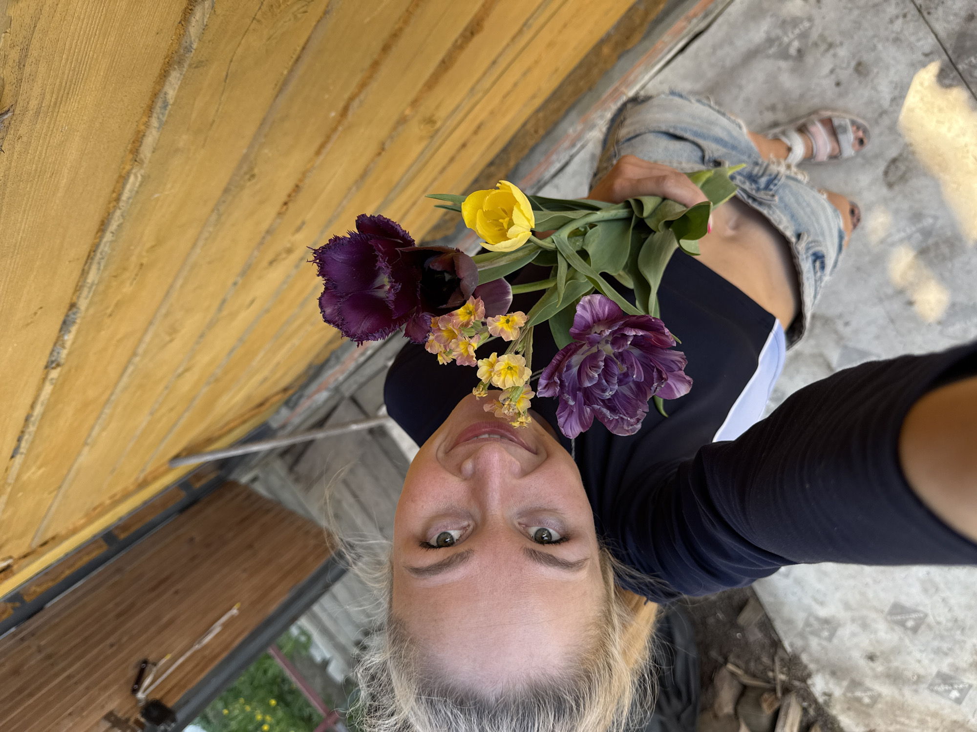 A woman takes a selfie while holding a bouquet of flowers. The bouquet contains at least one yellow tulip and two purple tulips, as well as some smaller yellow flowers. She is wearing a black top, denim shorts, and sandals. The background consists of a wooden wall or structure and a concrete floor. The location is Chaikovsky, Russia.