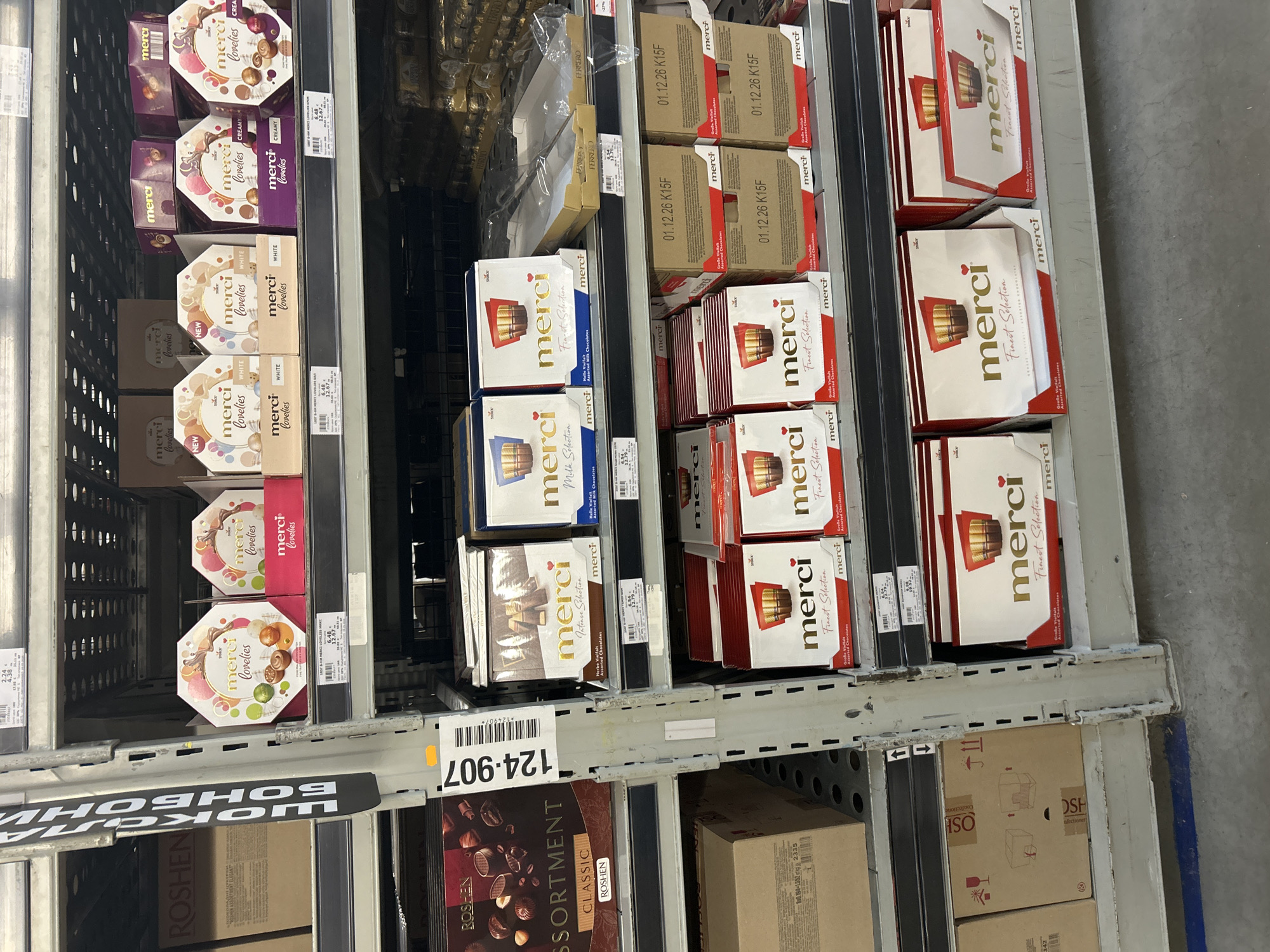 The image shows shelves stocked with Merci chocolates and Roshen assortments. The Merci chocolates come in varieties such as Finest Selection, Milk Selection, Intense Selection, and Lovelys in flavors like White and Creamy. Some of the Finest Selection boxes have the production date "01.12.26 K15F" printed on them. The product code "124-907" is also visible on the shelf. The shelves appear to be located in a retail environment, possibly a supermarket or wholesale store in Burgas, Bulgaria, due to the Cyrillic text visible on some labels and the presence of Roshen products.