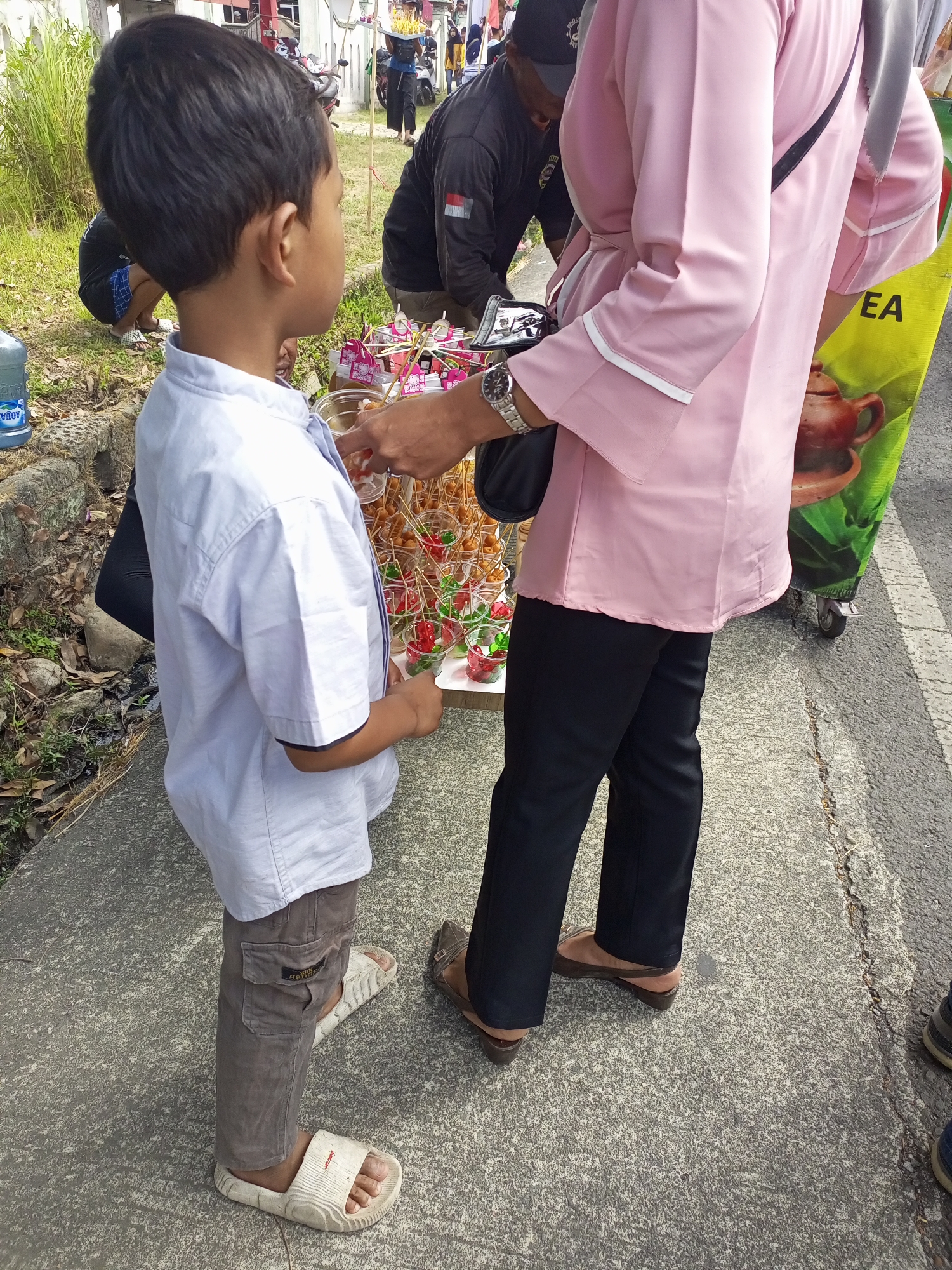 The image depicts a street scene in Nanggulan, Indonesia. A young boy wearing a light blue shirt and grey cargo pants stands on a concrete sidewalk, facing right. He is wearing white sandals. To his right, a person in a pink long-sleeved shirt and black pants is interacting with a display of food items, which appear to be skewers of small snacks in clear plastic cups with decorative toothpicks. The person is wearing brown wedge sandals and a watch on their left wrist. Behind the boy, a large blue water jug is visible, along with some foliage and rocks. In the background, other individuals and a cart are present, suggesting a market or street vendor environment. The lighting indicates it is daytime.