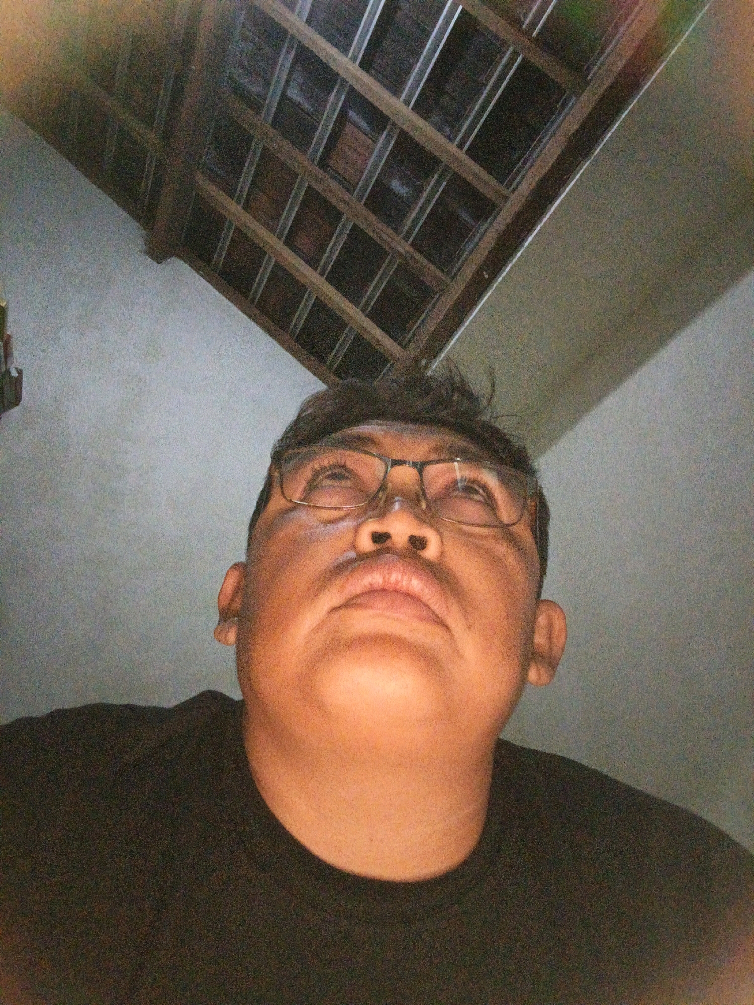 A low-angle, close-up, self-portrait photograph shows a man wearing glasses and a black t-shirt. He is looking upwards. The background consists of a textured wall and a wooden ceiling structure. The image was captured in Ketegan, Indonesia.