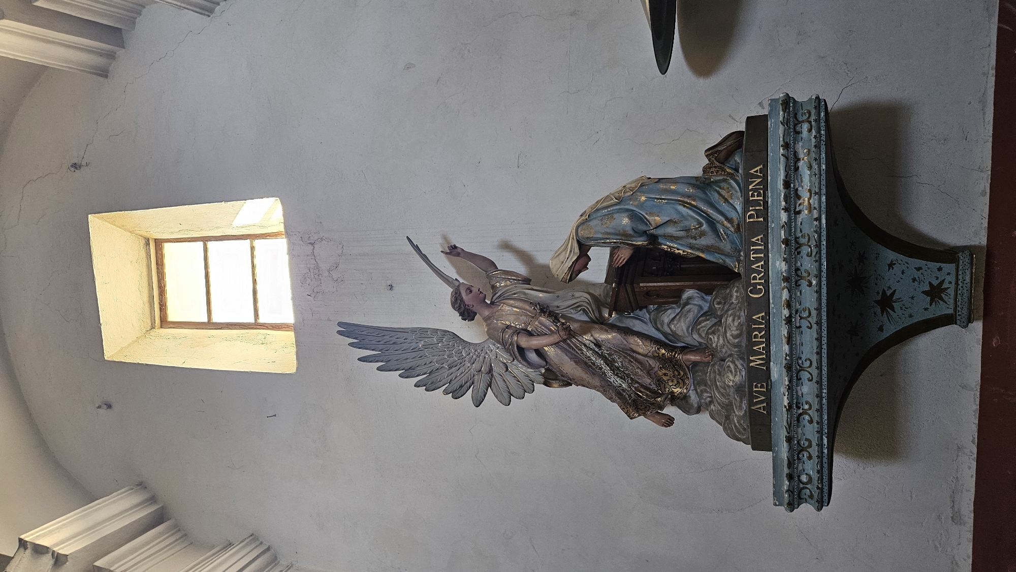 A statue depicting the Annunciation is displayed on a shelf in Sant'Antonino, France. The statue features an angel with outstretched arm and wings on the left, and a kneeling woman in blue robes on the right. Both figures are intricately detailed and colored, with the angel wearing a gold and white garment and the woman a blue and gold one. The shelf itself is ornate, painted in shades of blue and adorned with gold accents and star patterns. Above the statue, a simple rectangular window with wooden panes is visible, letting in a soft, diffused light. The wall behind the statue is textured and appears to be made of stucco, with some visible cracks. The overall setting suggests an interior space, possibly a church or a chapel, given the religious nature of the artwork and the architectural style.