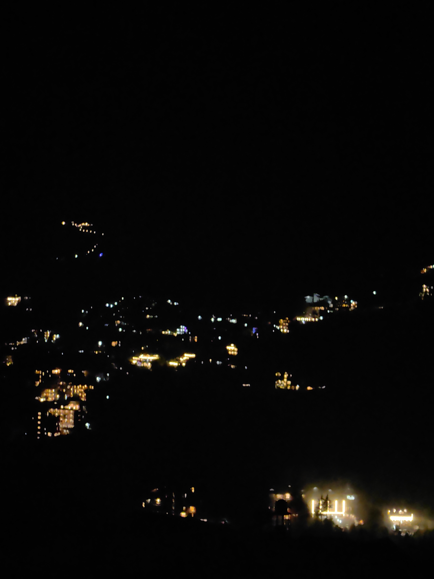 The image is a nighttime long-exposure photograph of a hillside illuminated by numerous lights from buildings and streetlights. The scene depicts a town or village nestled on a slope, with lights scattered across the dark landscape. Some clusters of lights suggest concentrated areas of habitation or commercial activity, possibly hotels or guesthouses. The lights vary in intensity and color, with many appearing as yellow or white orbs, while others have a more diffuse glow. The overall impression is of a settlement coming to life at night, with the lights creating a warm, inviting ambiance against the deep blackness of the night sky. The image captures a picturesque view of Vashisht, India, known for its hot springs and temples, suggesting a bustling atmosphere even after dark.