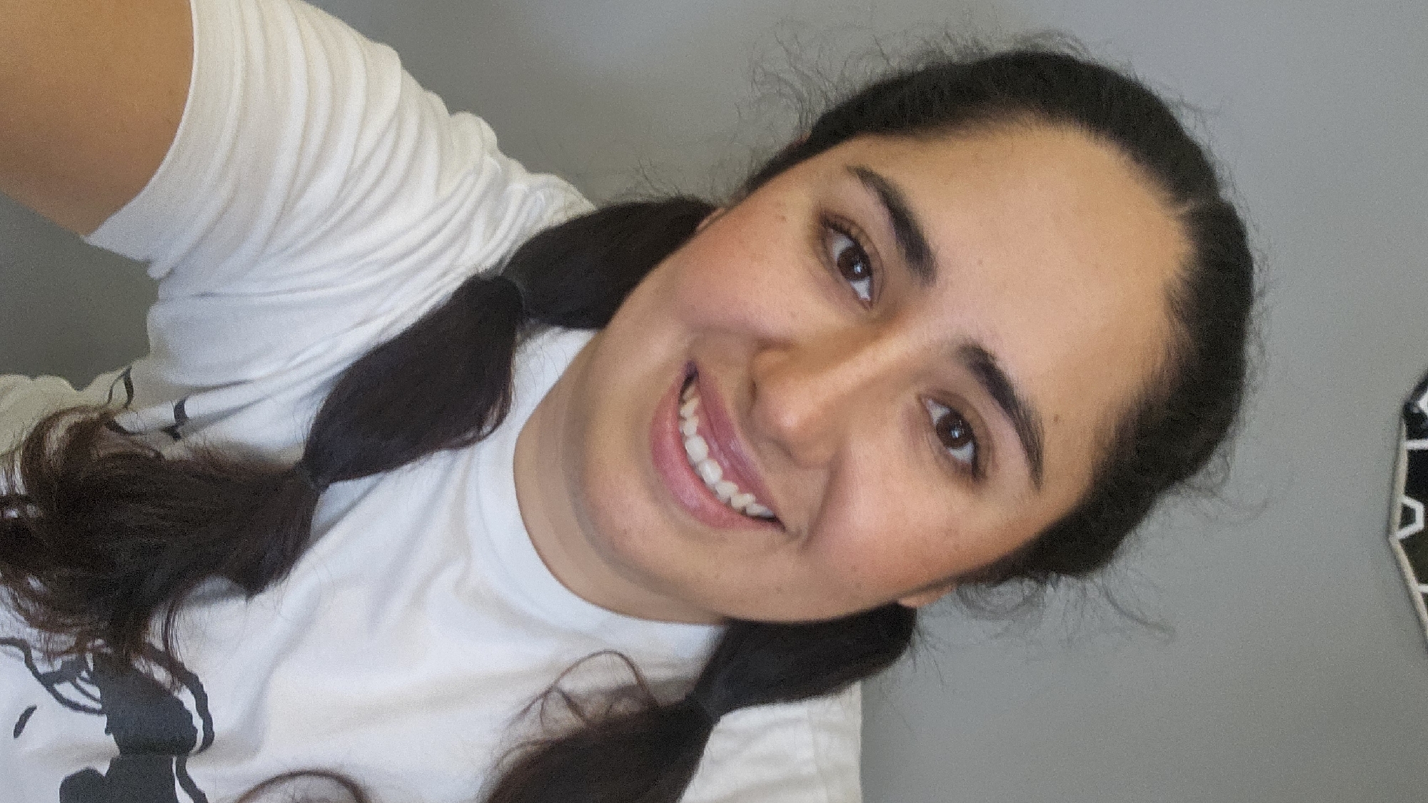 A young woman is captured in a close-up selfie, likely taken indoors in Puebla, Mexico. She is smiling broadly and looking directly at the camera, with her head slightly tilted to the side. Her dark, thick hair is styled into two long braids or pigtails that fall over her shoulders and chest. She has a bright, friendly expression, with a wide smile revealing her teeth. She is wearing a plain white t-shirt, on which a dark graphic design is partially visible towards the bottom left. This graphic appears to be a silhouette of a cartoon character, possibly resembling Mickey Mouse, with discernible ears and head shape. Her complexion is warm, with a hint of blush visible on her cheeks, and her eyes are dark brown. The background behind her is a simple, light gray wall, indicating a clean and uncluttered indoor setting. The lighting is bright and even, highlighting her features. No other discernible objects or text are clearly visible in the background.