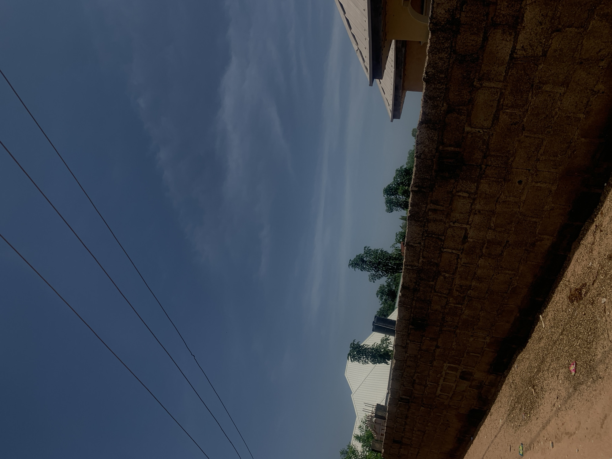 The image depicts a street scene in Jalingo, Nigeria, under a partly cloudy sky. In the foreground, a dirt road curves towards a rough, tan-colored brick wall that stretches across the frame. The wall appears aged and weathered, with visible mortar lines and some signs of wear. Beyond the wall, several buildings are partially visible. To the left, a white, angular roof with a corrugated metal surface suggests a modern construction, possibly a warehouse or large home. Several trees with dark green foliage are interspersed between the buildings. To the right, a portion of another building with a light brown, sloped roof and yellow-colored walls is visible. The sky above is a deep blue, with streaks of lighter clouds, and three parallel dark power lines are strung across the upper portion of the image. The overall scene suggests a residential or semi-industrial area on the outskirts of the city.