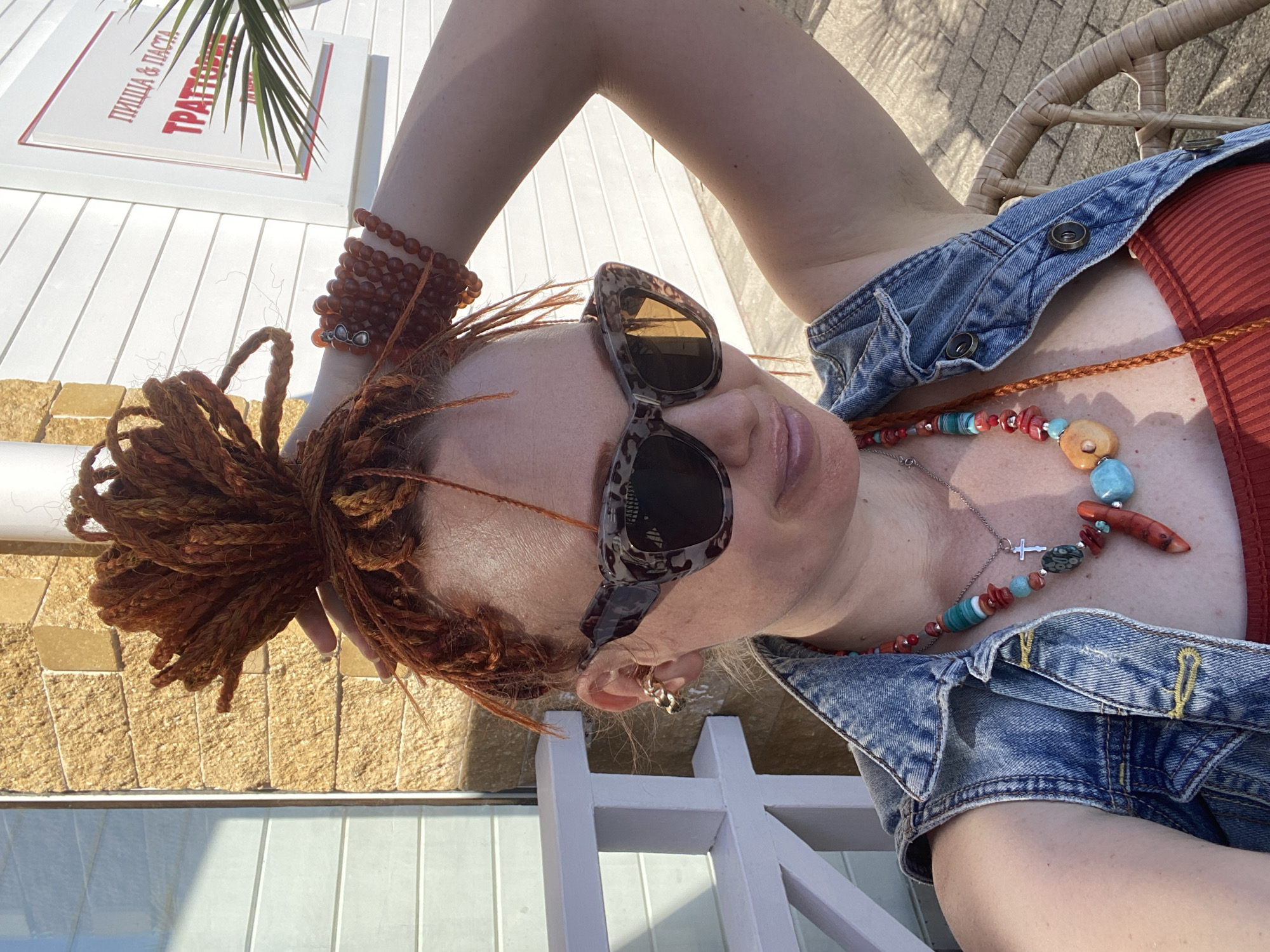 The image shows a woman with red dreadlocks and a denim vest posing outdoors. She is wearing sunglasses with a leopard print frame and a colorful beaded necklace with a cross pendant. Behind her, a white building with a sign that reads "PIZZA & PASTA TRATTORIA" is visible. A palm frond is also present in the background. The scene appears to be in a sunny outdoor setting, likely a cafe or restaurant patio, which aligns with the typical atmosphere found in Sochi, Russia, a city known for its resorts and coastal attractions. The woman's posture and the overall lighting suggest a warm day.