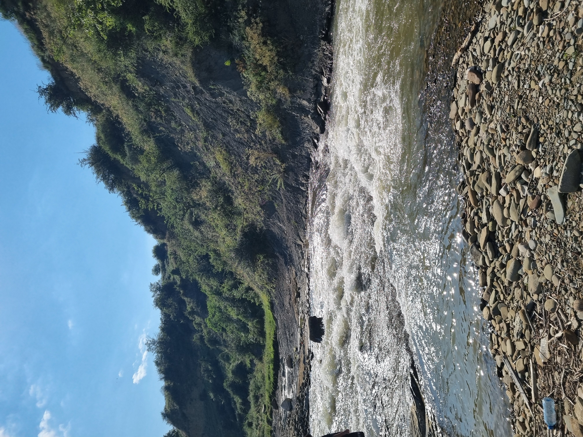 A wide shot captures a sun-drenched, natural scene in Khuchni, Russia, featuring a shallow, fast-flowing river with a rocky bed and banks. The water is turbulent, with white rapids breaking over submerged stones, reflecting the bright sunlight. The left bank of the river is covered in pebbles and driftwood, with a lone plastic bottle visible near the bottom left. A person, partially visible from the waist down, stands in the shallows on the far left. The right bank is a steep, eroded hillside covered in dense green vegetation and trees, with patches of bare earth. The sky above is a clear, bright blue with a few wispy clouds.