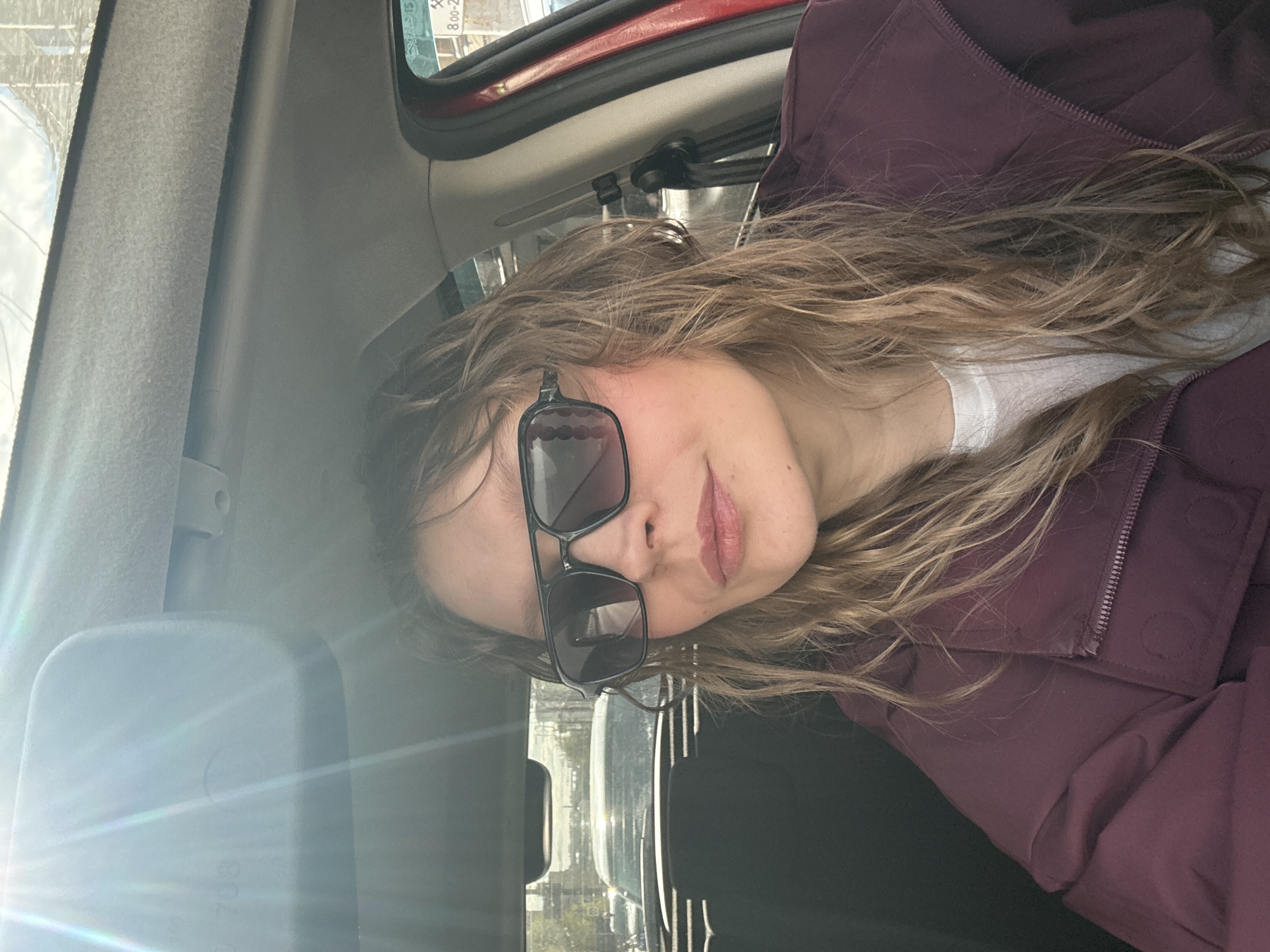 A woman wearing sunglasses and a burgundy jacket is sitting in a vehicle. The interior of the vehicle is visible, including the dashboard and parts of the windshield. Outside the vehicle, a city street scene is visible, with other cars and buildings. The location is Izhevsk, Russia.