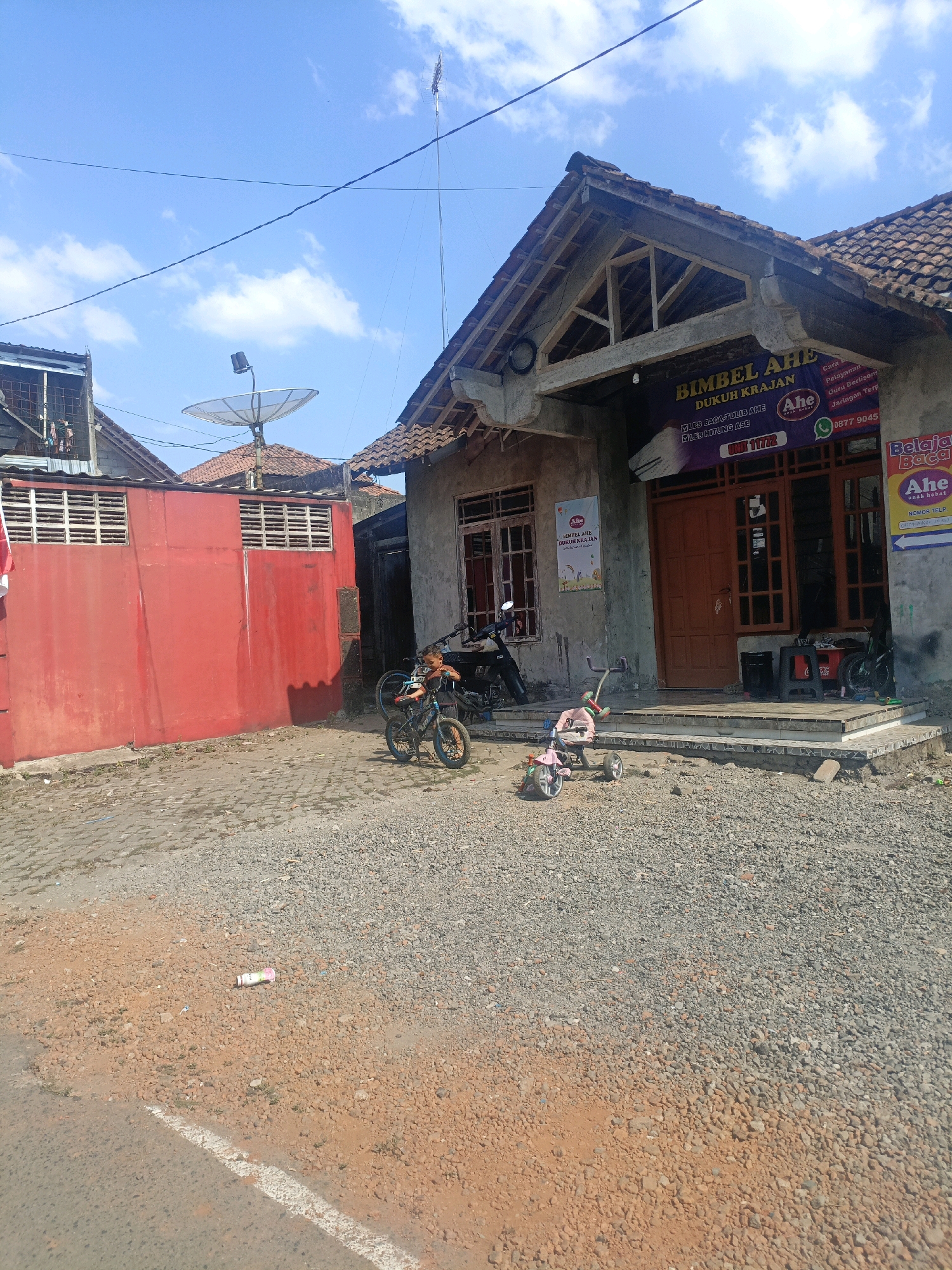 The image depicts a street scene in Salatiga, Indonesia, on a sunny day with a bright blue sky and scattered clouds. In the foreground, a gravelly dirt road with a white painted line runs diagonally across the bottom. Scattered on the road are small rocks and some reddish-brown earth. A pink and white bottle lies on its side in the gravel. Further back, a paved area with stones is visible.
Dominating the mid-ground and background is a building with a tiled roof. A sign above the entrance reads "BIMBEL AHE DUKUH KRAJAN" and other text in Indonesian, indicating a tutoring center. The building's facade appears weathered and unfinished in some areas, with exposed wooden beams visible under the eaves. Orange-brown double doors serve as the entrance. To the left of the building is a bright red wall with horizontal white slats, characteristic of a fence or boundary.
In front of the building and extending towards the red wall, there are several objects. Two children's bicycles, one blue and black with training wheels, and another blue bicycle, are parked near the entrance. A pink tricycle with a yellow handlebar is positioned in front of the bicycles. A person is partially visible near the blue bicycle, appearing to be a young boy. A black motorcycle is parked on the steps leading to the building.
Various satellite dishes and antennas are visible on rooftops in the background, along with more residential structures. The overall scene conveys a sense of a bustling, yet somewhat rustic, urban environment.
