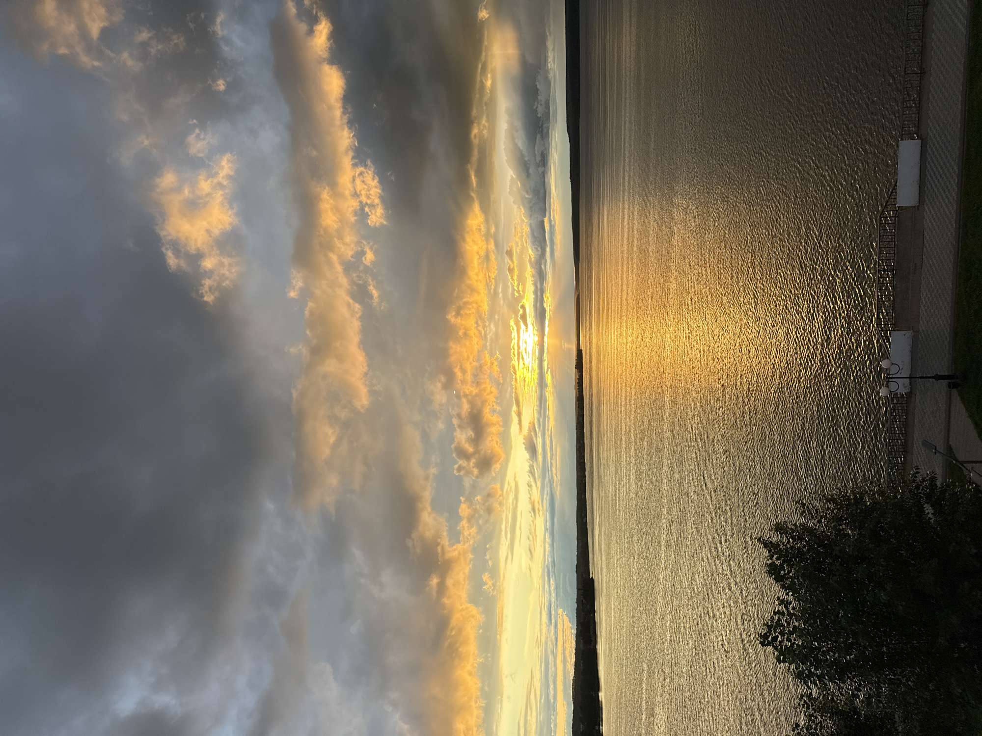 This media file depicts a serene sunset over a wide body of water, likely a river or lake, with a backdrop of dramatic clouds. The sun is low on the horizon, casting a golden, shimmering reflection across the rippling surface of the water, which occupies the right two-thirds of the frame. The water's texture is characterized by numerous small waves, creating a pattern of light and shadow.
On the far right, a paved walkway with a decorative railing and a lamppost is visible, suggesting a public promenade or embankment. A dark silhouette of a leafy tree is prominent in the lower right corner, partially obscuring the view of the walkway and the water's edge. The left third of the image is dominated by the sky. The clouds are a mix of dark, brooding grays and lighter, wispy formations tinged with orange and yellow from the setting sun. The light breaks through the clouds in several places, creating beams that illuminate the sky and add depth to the scene.
The overall mood is peaceful and contemplative, with the vibrant colors of the sunset contrasting with the darker, more subdued tones of the clouds and foreground foliage. The time of day is clearly sunset, and the weather appears to be fair despite the presence of clouds. While no people are visible, the presence of the walkway and lamppost implies a populated area. There is no visible text in the image. The location context provided is Votkinsk, Russia, which aligns with the visual cues of a wide river or lake and a developed embankment.