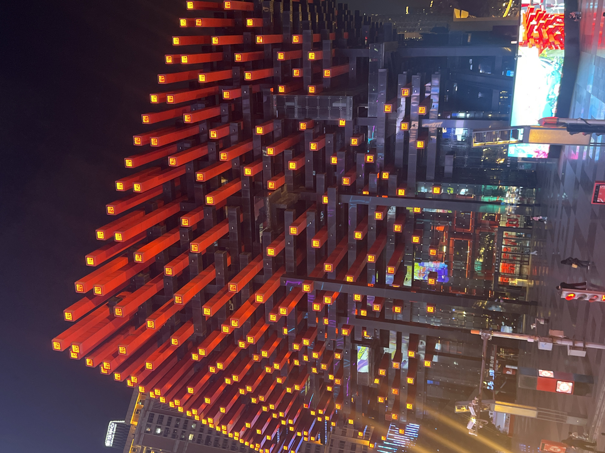 The image depicts the Hongyadong complex in Yuzhong District, Chongqing, China, at night. The structure features stacked, dark-colored supports from which extend numerous horizontal red beams, each adorned with a yellow square emblem. The complex abuts a street with visible pedestrian traffic signals and a large screen displaying bright, colorful images. Adjacent buildings with illuminated windows and streetlights are also visible.