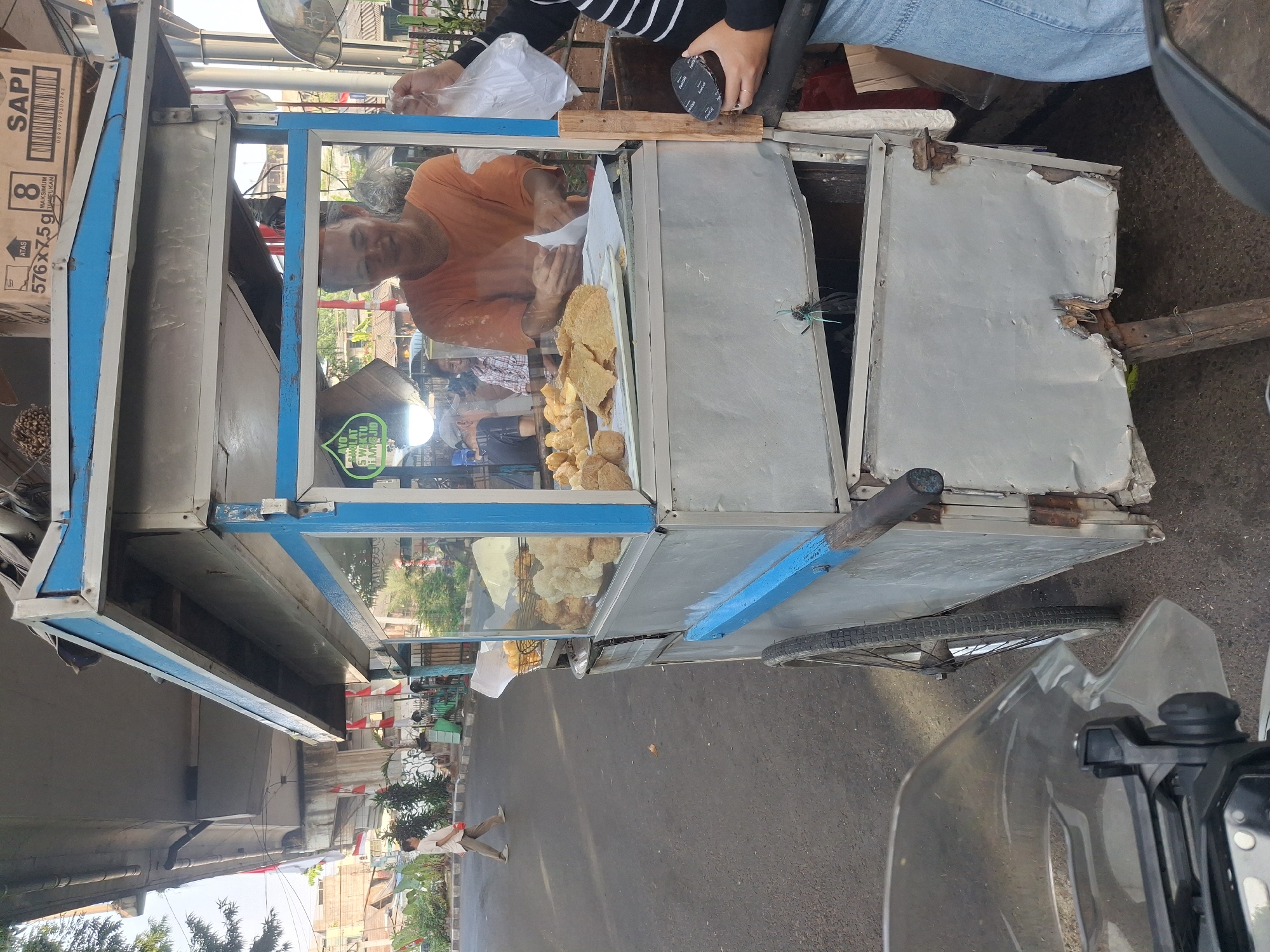 This is a medium shot taken from a motorcycle, looking towards a food cart under an overpass in South Jakarta, Indonesia. The food cart is constructed from metal and has blue accents. A man in an orange shirt is serving food from behind a glass display case filled with fried snacks. A person is holding a plastic bag and a small dark container. In the background, a man in a white shirt walks past on the sidewalk. Red and white Indonesian flags are visible on the overpass. The overall scene depicts a street food vendor in an urban environment.