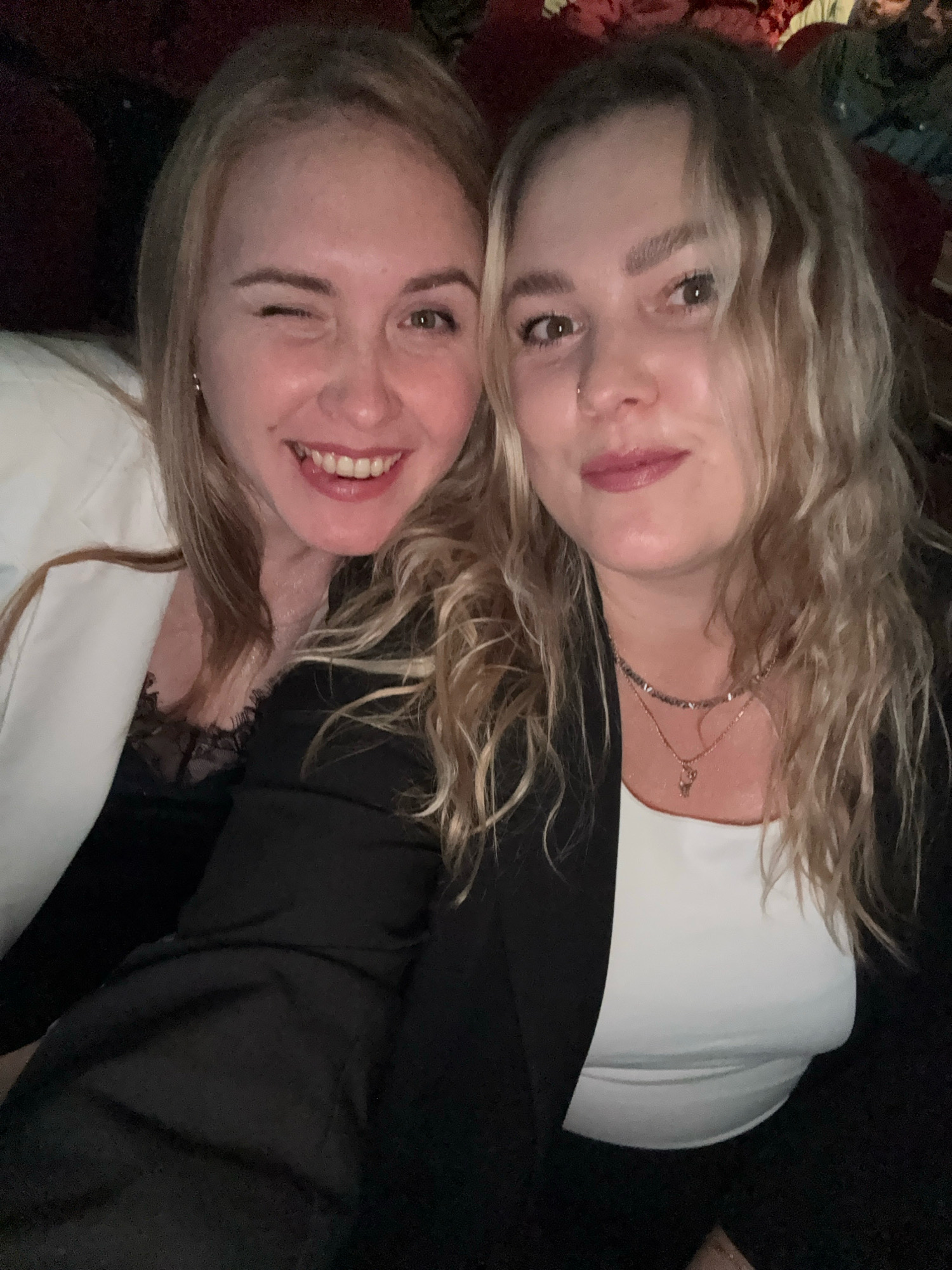 Two blonde women are taking a selfie. The woman on the left is winking and smiling. She is wearing a white blazer over a black lace top. The woman on the right has wavy blonde hair and is smiling. She is wearing a black blazer over a white top and a necklace. The background is dark and appears to be an indoor setting, possibly a dimly lit room or a cinema. There are no visible landmarks or context to suggest the location is in Марковский, Russia.