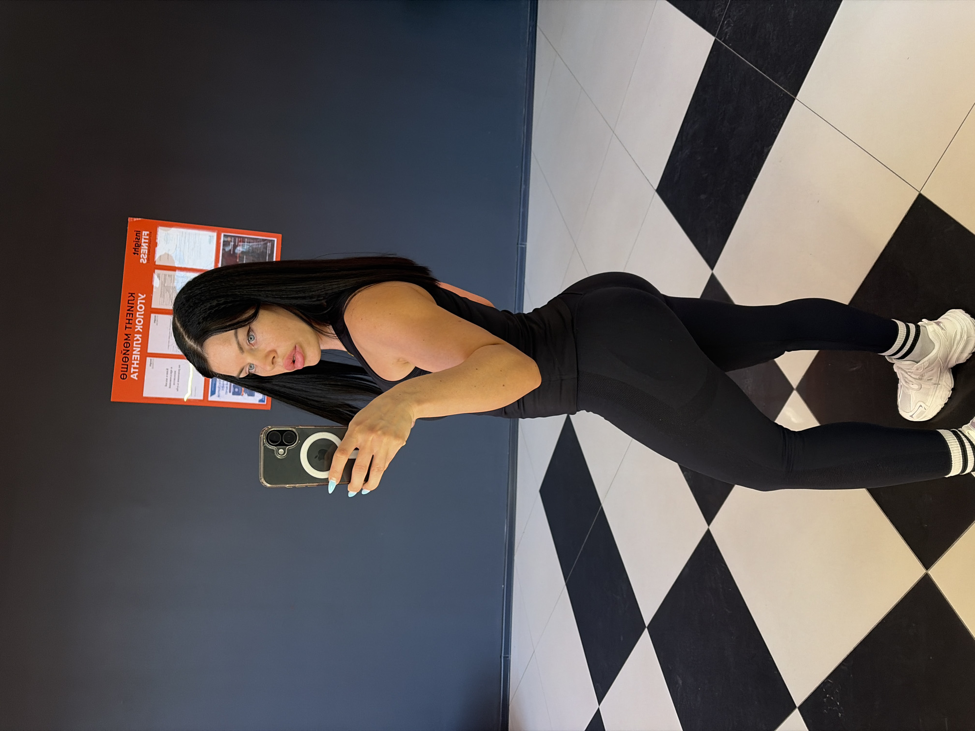 In a gym with a black and white checkered floor and dark grey walls, a woman with long black hair is taking a selfie. She is wearing black athletic leggings, a black tank top, and white athletic shoes with black and white striped socks. She is holding a smartphone in her left hand, and her right arm is bent at the elbow. A red sign with white text is visible on the wall behind her. The sign appears to be in Russian and contains information about fitness. This scene is in the city of Ufa, Russia.