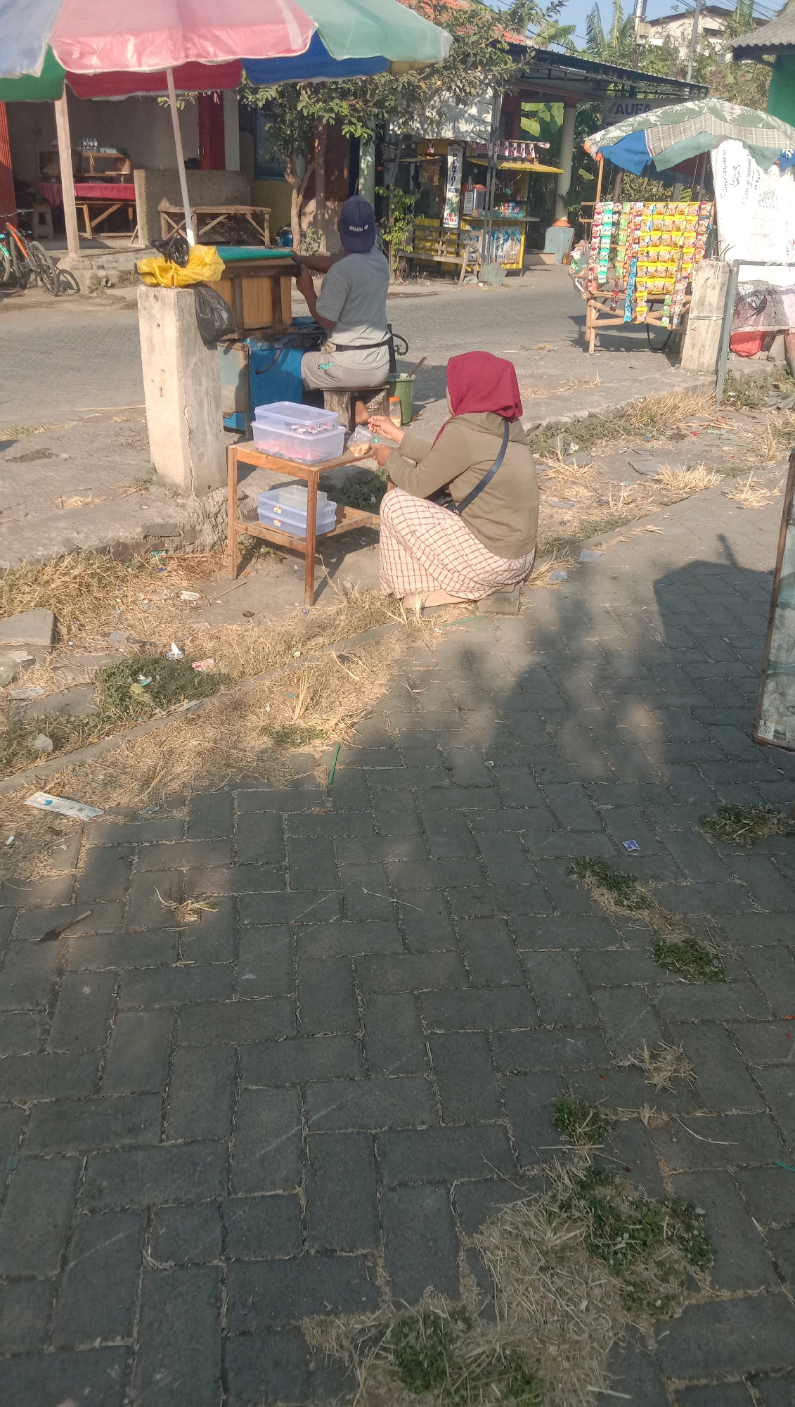 The image captures an outdoor scene on a sunny day, likely in Curah Dukuh, Indonesia. In the foreground, a person wearing a red hijab and a khaki jacket is seated on the ground, facing away from the camera and towards a small wooden stand. They are holding a piece of food in their hands, possibly eating. The stand has two shelves, with clear plastic containers on the lower shelf and another container on the upper shelf. To the left of the stand, another person wearing a grey t-shirt and a dark cap is seated on a wooden stool. They are also facing away from the camera and appear to be engaged in some activity at a workbench. A yellow plastic bag and a black trash bag are visible on a concrete structure behind the workbench. In the background, there are several small stalls or carts selling various goods, including packaged snacks. The ground is paved with dark gray interlocking bricks, and dry grass and debris are scattered around. A pink, red, and green umbrella is set up over the workbench area. The overall impression is of a street-side vendor setup, likely a food stall.