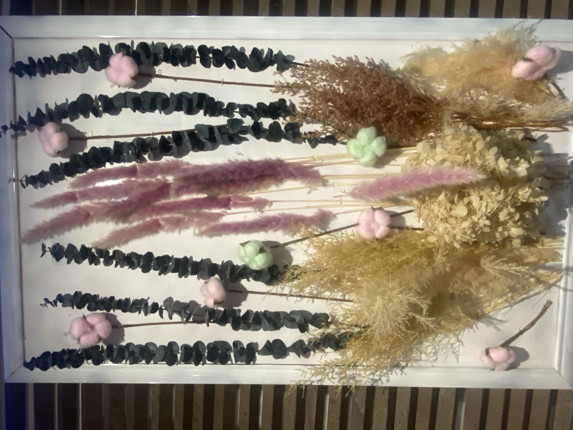 A framed arrangement of dried flowers and eucalyptus branches is displayed on a wall. The arrangement features a variety of textures and colors, including pink feathery plumes, tan grasses, and dark eucalyptus leaves. Small, cotton-like embellishments in pink and light green are interspersed throughout the arrangement. The background of the image appears to be a wall with horizontal wood paneling, suggesting an indoor setting. The specific location of this arrangement in Ivanteyevka, Russia, cannot be determined from the provided image. There are no people or actions depicted in the scene.
