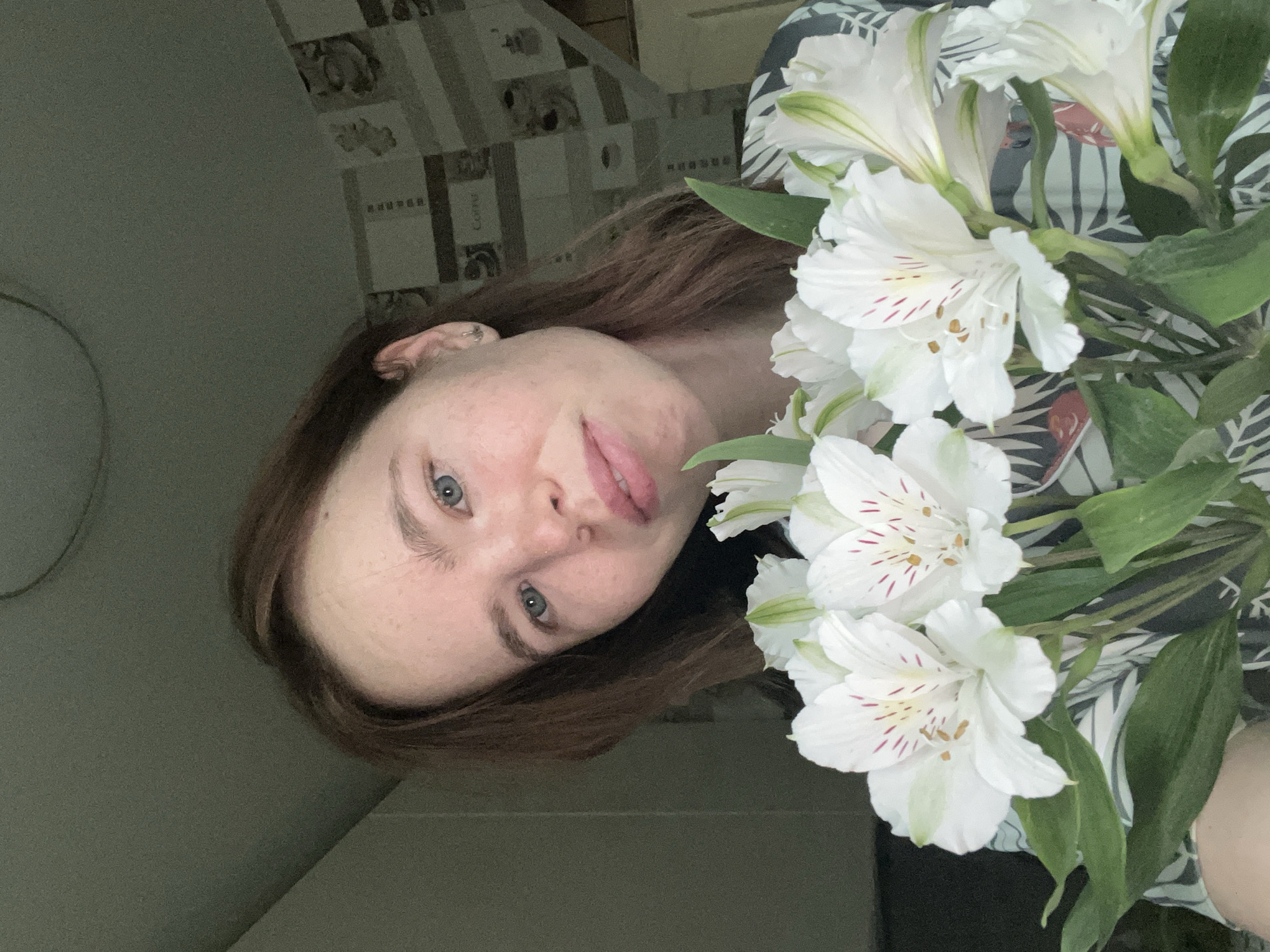 The image shows a young woman with brown hair and blue eyes holding a bouquet of white alstroemeria flowers. She is wearing a patterned shirt. The background appears to be an indoor setting with muted colors and a patterned wallpaper. The lighting suggests it is daytime. The location is identified as Chaikovsky, Russia, though specific landmarks are not visible.