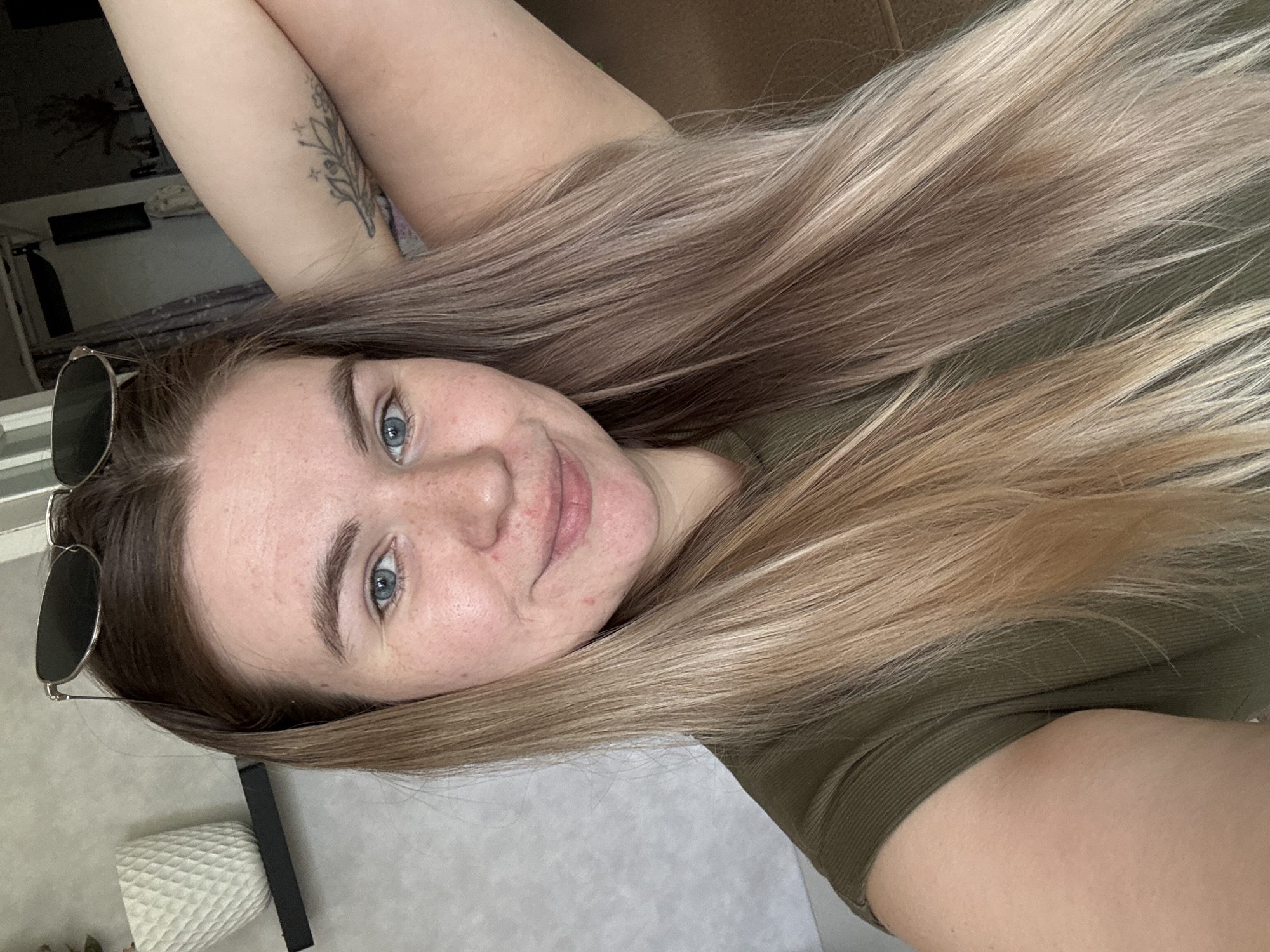 A woman with long, blonde and brown hair is visible in a close-up, selfie-style photograph. She has fair skin, blue eyes, and visible freckles. She is wearing a green, ribbed shirt. Sunglasses are perched on her head. A tattoo is visible on her upper left arm. The background includes a glimpse of a room interior, possibly a bedroom, with a dark-colored object near a wall, and a partially visible white object that could be a lampshade. This image cannot be definitively tied to the city of Chaikovsky, Russia based solely on the visual information provided.