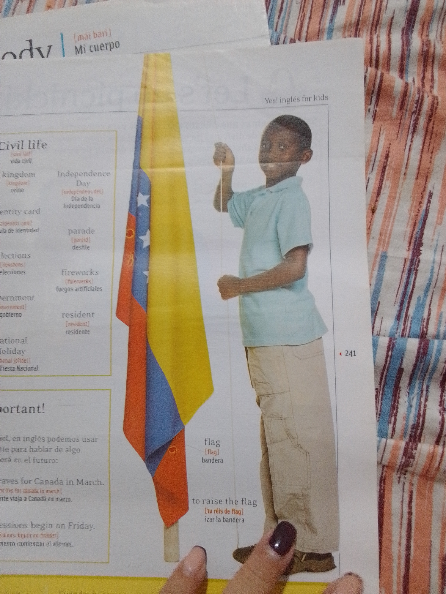The image shows a page from a bilingual English-Spanish children's book. The page is divided into columns of text and an image of a young boy holding a Colombian flag. The text appears to be vocabulary related to civic life, with English words and their Spanish translations. The boy is wearing a light blue polo shirt and tan pants. The flag is prominently displayed, with its yellow, blue, and red stripes visible. The background is a striped fabric, possibly a blanket or tablecloth. The image is cropped to focus on the boy and the flag, with the text on the left side of the page. There is a page number "241" on the right side. No location in the city of La Tagua, Venezuela is depicted.