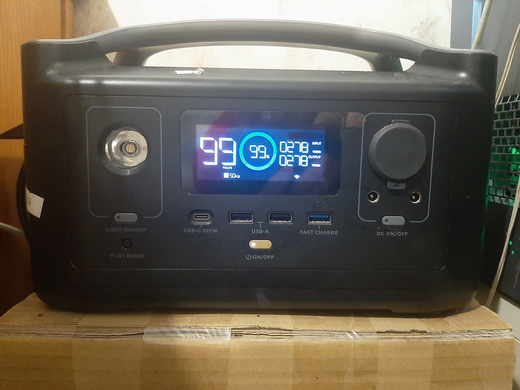 An outdoor power station is shown sitting on top of a cardboard box. The power station is black and has a digital display screen that shows "99" for hours, a circular icon with "99%" inside, and "0278 Watts" for both input and output. There are various ports on the front of the device, including a USB-C port, USB-A ports, and DC ports. A circular knob is also visible on the right side of the device. The environment appears to be indoors, with a wooden surface in the background and what looks like a computer tower to the right. The lighting is artificial.