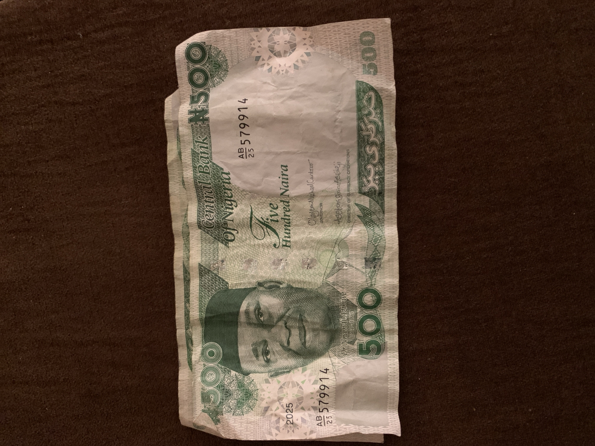 This is a close-up, top-down shot of a Nigerian 500 Naira banknote, lying flat on a brown, textured surface. The banknote is slightly creased and wrinkled, with the green design and portrait of Dr. Nnamdi Azikiwe prominently visible. The denomination "500" is repeated in large green numerals on the left and right sides of the bill, as well as in the center. Text on the banknote includes "Central Bank of Nigeria," "Five Hundred Naira," signatures of "Olayemi Michael Cardoso" (Governor) and "Adetona S. Adefideji" (Director of Currency Operations), and a serial number "AB 579914." A date "2025" is also visible. There is no indication of the city of Jos, Nigeria, in the image.