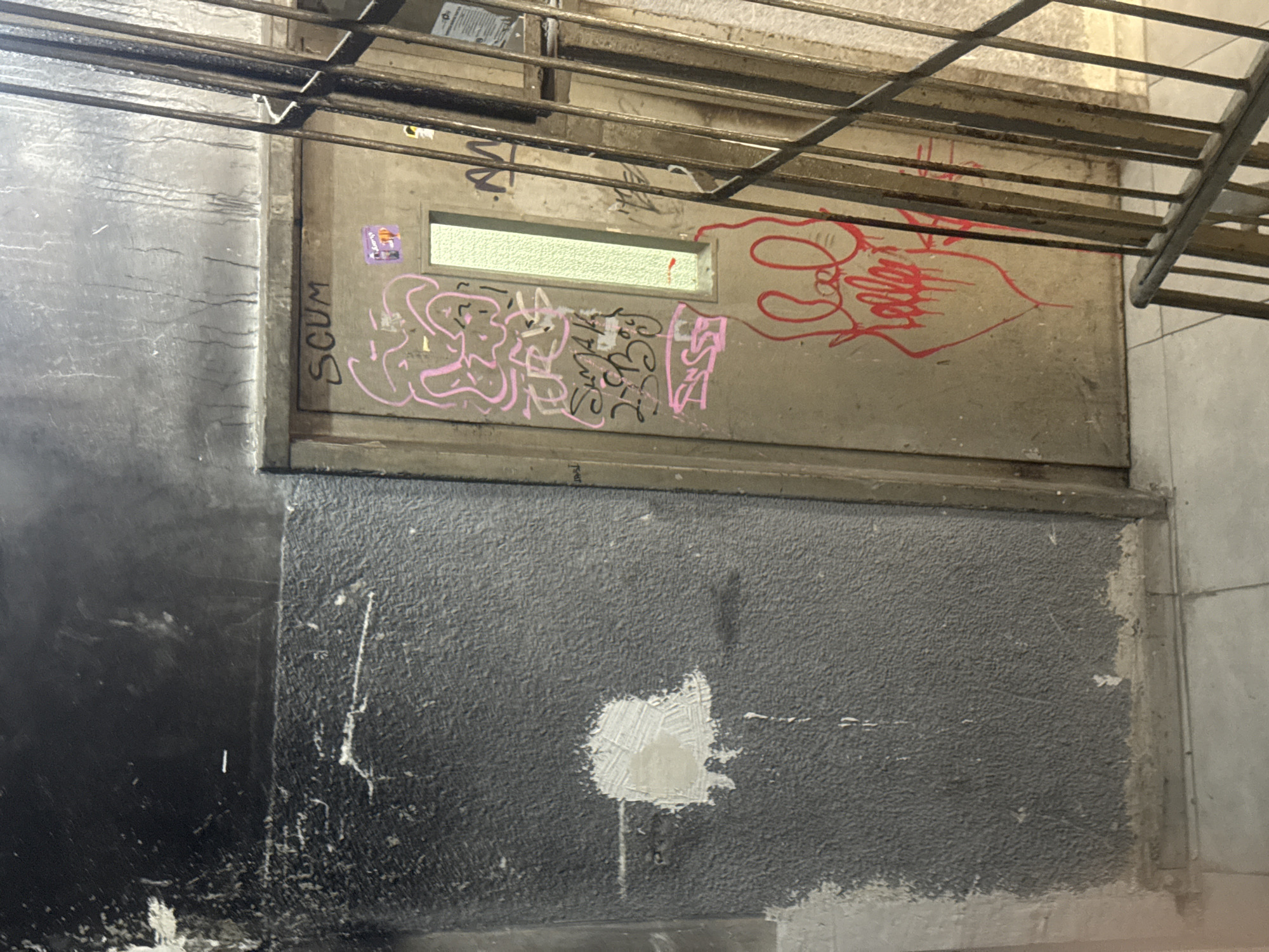 An exterior wall in an urban setting, possibly Tel Aviv, Israel, featuring a utility access point or hatch. The upper section is a gray, weathered panel with graffiti in pink and red paint, including illegible tags and the word "SCUM." A horizontal window or opening is located near the top of the panel. The lower section of the wall is a rough, dark gray surface with a patch of light-colored filler or repair material. A metal grid or grating structure is visible above the upper panel, suggesting an overhead walkway or support. The overall scene conveys neglect and decay, with signs of vandalism and deferred maintenance.