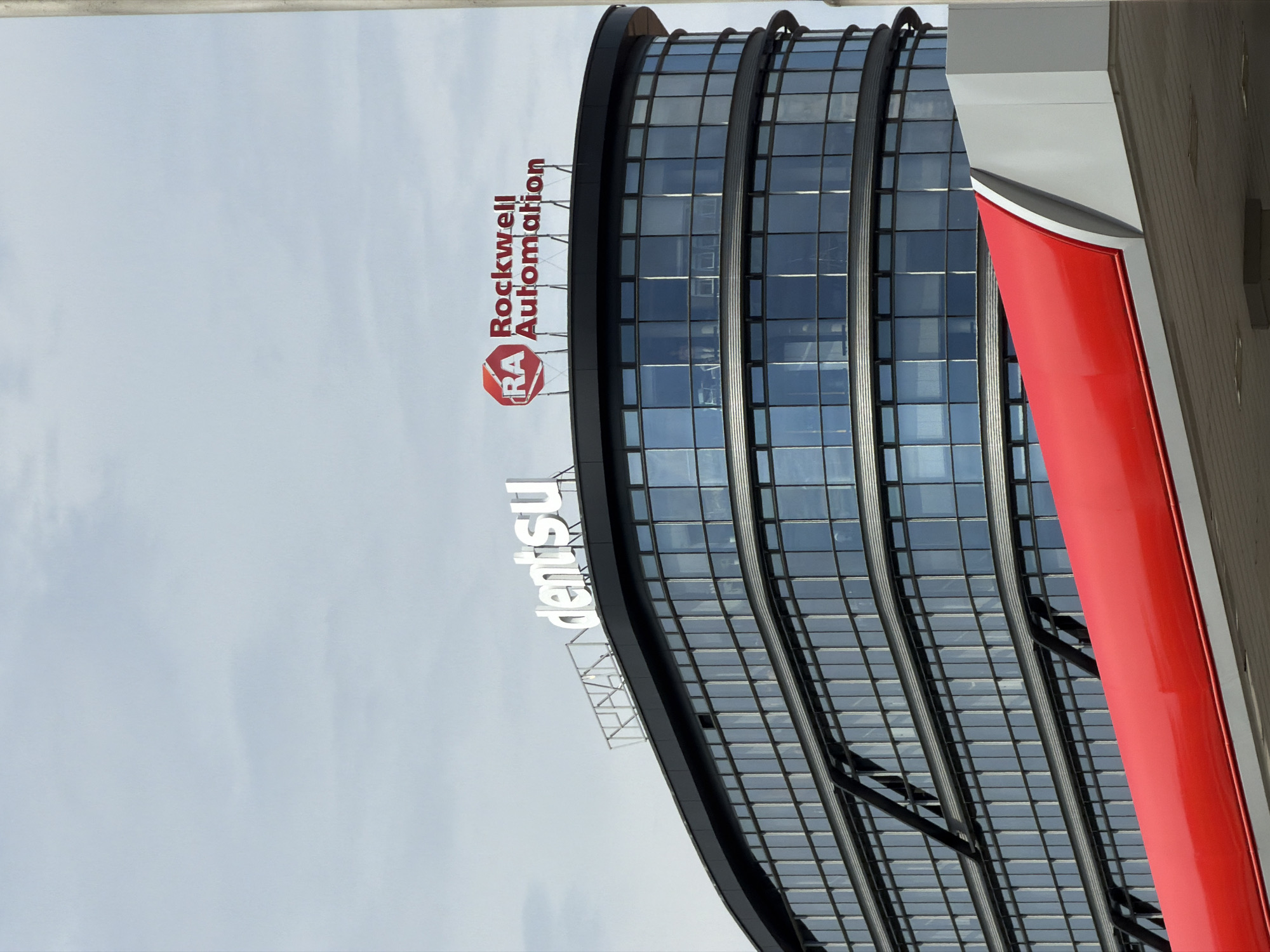An outdoor, eye-level, slightly low-angle, full-shot photograph captures the upper floors of a modern, curved office building situated in Prague, Czechia. The building's exterior is characterized by a sleek, dark facade with multiple rows of large, reflective glass windows. Some of these windows are accented by metallic horizontal louvers or awnings that extend outwards.
Prominently displayed on the rooftop of the building are two distinct signs. The left sign features white, illuminated, three-dimensional lettering spelling out "dentsu". To the right of the "dentsu" sign, a second logo and text are visible. This consists of a red and white circular emblem with the letters "RA" inside, followed by red, illuminated, three-dimensional lettering that reads "Rockwell Automation".
In the foreground, a portion of a structure with a bright red, curved canopy or overhang is visible, extending into the bottom of the frame. This red element is partially obscured by the bottom edge of the image, creating a framing effect. The sky above is overcast with a pale, cloudy texture. No people are present in the scene. The overall impression is one of corporate architecture and branding against a muted sky.