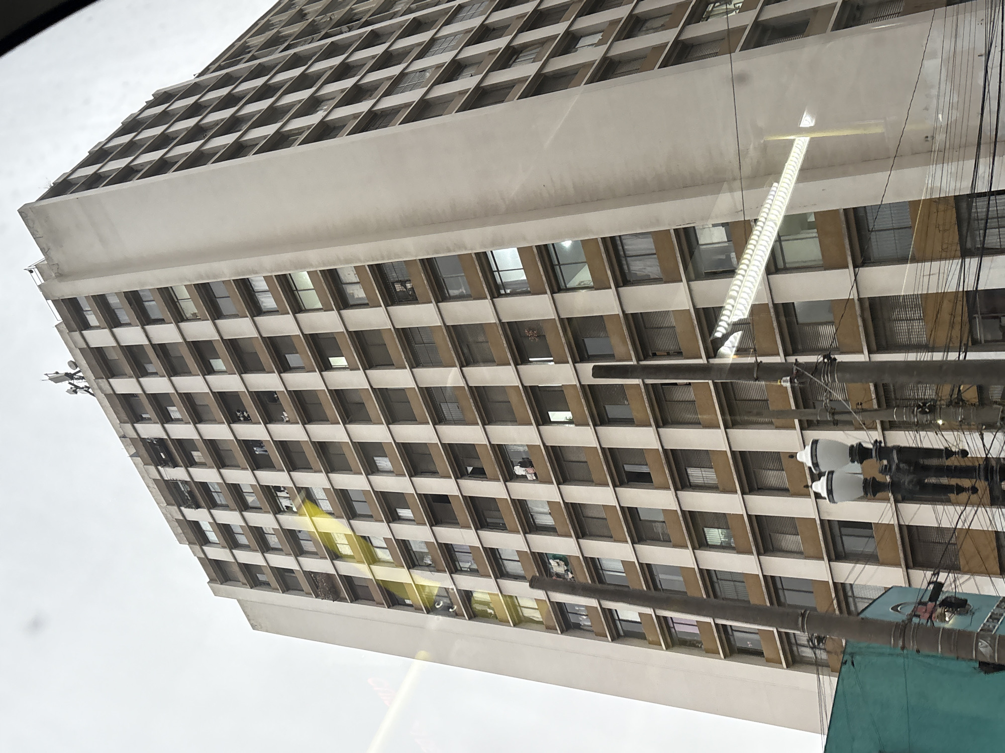 The image shows a tall, concrete apartment building with many windows, located in the city of São Paulo, Brazil. The building has a stark, utilitarian design with alternating bands of windows and beige concrete. The sky is overcast. There are several utility poles and wires in the foreground, as well as a street lamp. Reflections of lights and possibly other buildings are visible on the windows of the apartment building. Some laundry is hanging from balconies on several floors. The perspective is from below, looking up at the building.