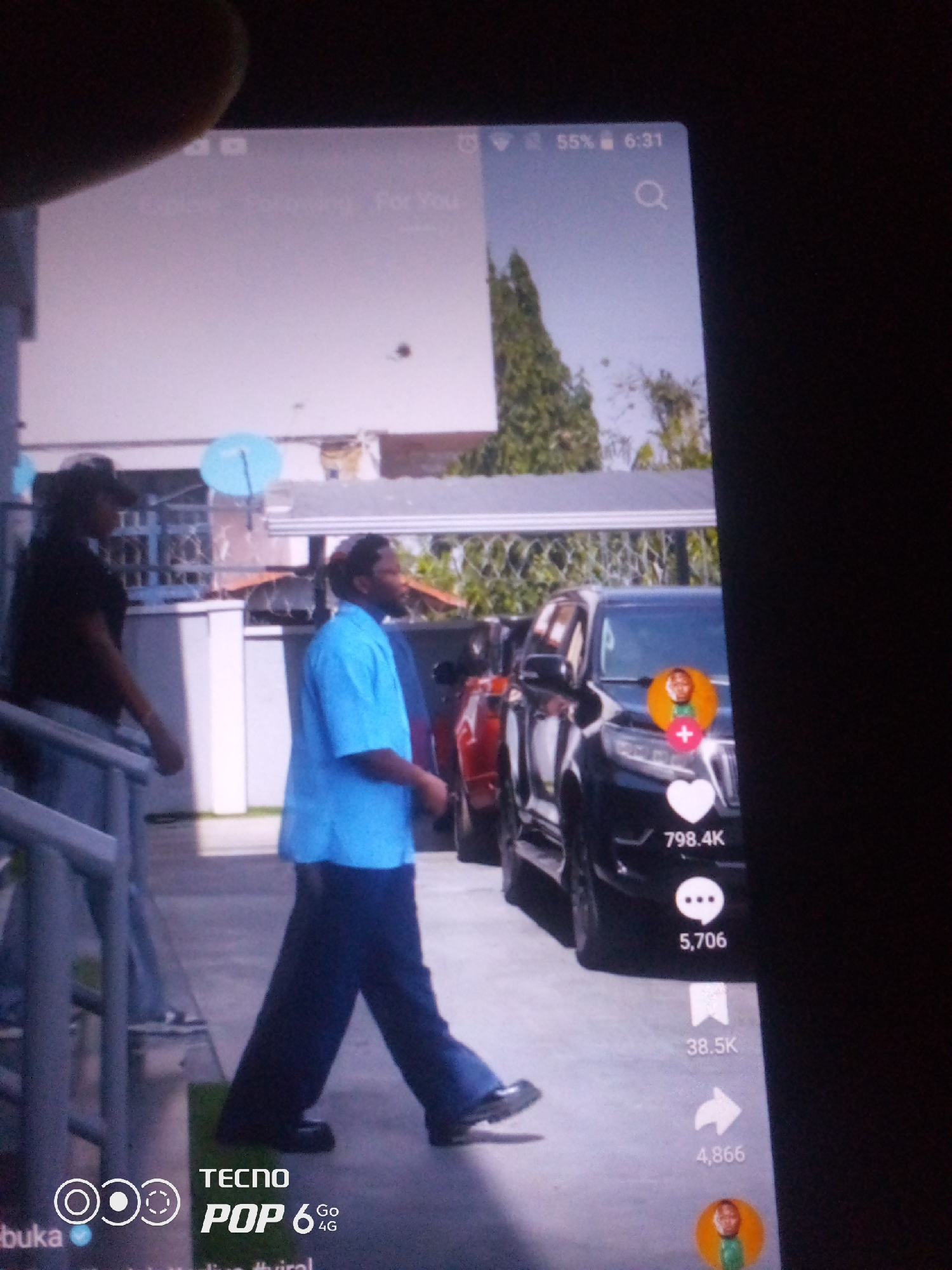 The image shows a close-up of a smartphone screen displaying a video. The video appears to be a social media post, possibly from TikTok, given the interface elements like likes, comments, shares, and a user profile icon. In the video, two individuals are visible. The person in the foreground is a man wearing a bright blue collared shirt and dark pants, walking with his body angled away from the camera. Behind him, a person in a black t-shirt and a cap is also visible, walking. They are outdoors, with a building and a parked black SUV in the background. The phone's display also shows status icons at the top, including battery level and time. The text overlay at the bottom of the screen indicates "TECNO POP 6 GO 4G." The overall scene is a street or outdoor area, likely in a city. There is no specific information in the image that would place the location in Howayi, Nigeria.