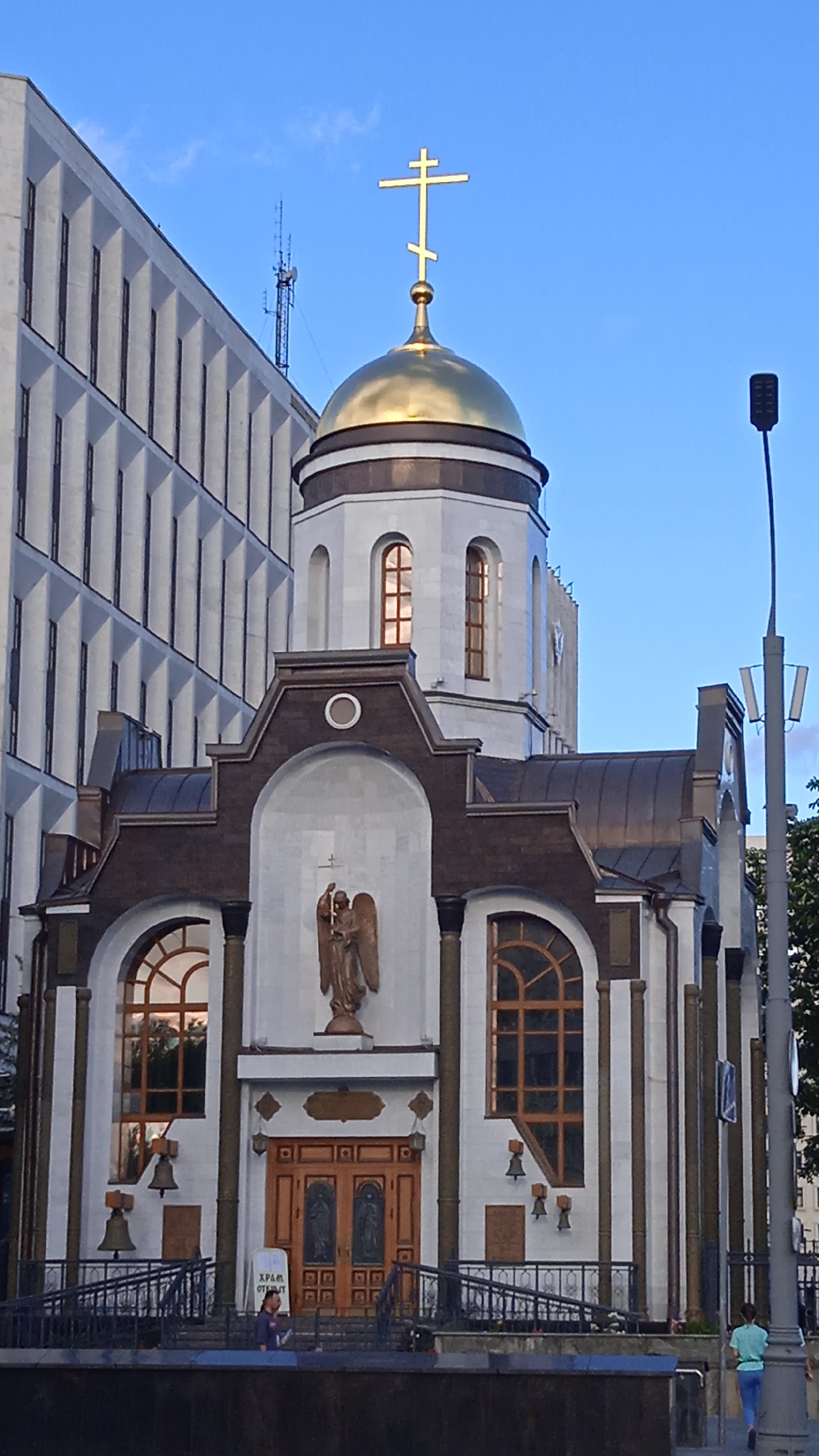 The image shows the Church of the Resurrection of Christ in Moscow, Russia, situated next to a modern building. The church features a golden dome topped with a triple-barred cross. The facade is white with brown architectural accents and features a statue of an angel above a set of double wooden doors. Several arched windows with wooden frames are visible, along with bells mounted on the exterior. In front of the church, there is a low fence and a set of stairs leading up to the entrance. Two individuals are present: one person is standing on the steps near the entrance, and another person is walking away from the church on the sidewalk to the right. A lamppost is visible to the right of the church. The sky is clear and blue.