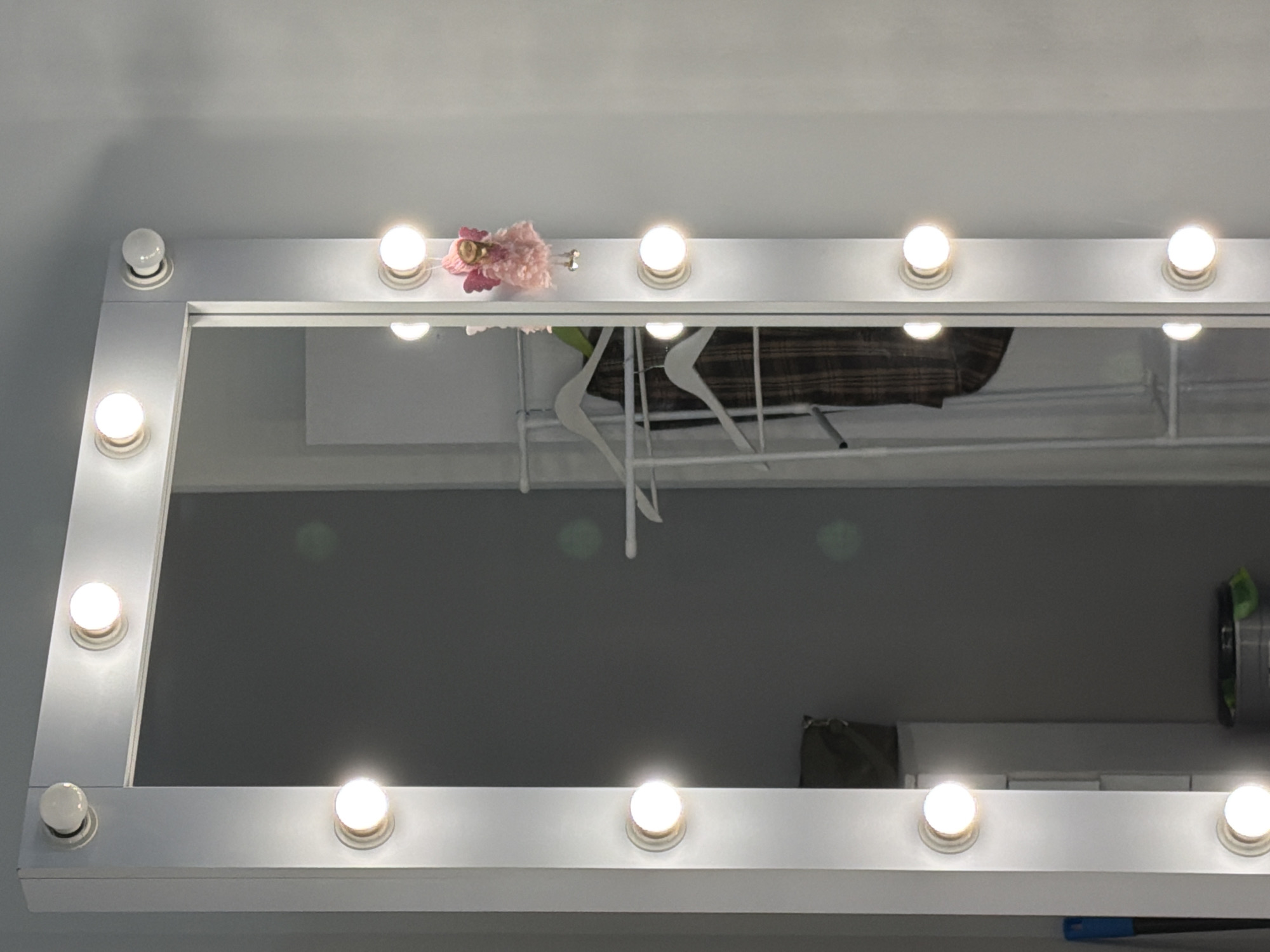 This media file features a close-up shot of a well-lit mirror, likely a vanity mirror with Hollywood-style bulbs. The mirror has a white frame and is adorned with several round light bulbs, some of which are illuminated. A small, pink, doll-like ornament with floral elements is attached to the top edge of the mirror frame.
In the reflection of the mirror, a clothes rack is visible, holding several white hangers. A dark, plaid garment is draped over one of the hangers, and a portion of a green item, possibly a bag or container, can be seen on the right side of the reflection. The background in the reflection appears to be a wall, suggesting an indoor setting.
The lighting in the image is bright and even, originating from the bulbs on the mirror. This suggests that the photo was taken in a well-lit indoor environment, possibly during the daytime or in a room with artificial lighting. There is no visible text in the image. The setting cues, such as the vanity mirror and clothes rack, indicate a dressing area or a personal space. The overall mood conveyed is neutral, focusing on the objects and their arrangement. The location context provided is Kyiv, Ukraine.
