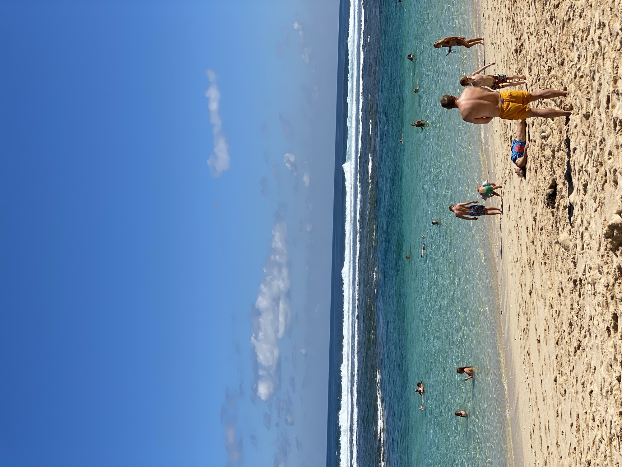 The image depicts a bright, sunny day at a beach in Kuta Selatan, Indonesia. The scene is dominated by a clear blue sky and a sandy beach that transitions into the turquoise waters of the ocean. A series of white waves are visible in the distance, suggesting a reef break or a strong surf.
Several people are scattered across the beach and in the shallow waters. On the sand, a man in yellow swim trunks stands with his back to the camera, looking out towards the ocean. Another person lies on the sand wearing patterned swim trunks. A child in green shorts is seen near the water's edge, and another person is lying on their back further up the beach. In the water, multiple individuals are seen swimming, wading, and relaxing. Some appear to be standing or floating, enjoying the clear, shallow sea.
The sunlight is bright, casting shadows on the sand and illuminating the water, highlighting its transparency. The overall mood is one of leisure and enjoyment, typical of a beach vacation. The presence of the ocean waves and the clear water strongly indicate a tropical or subtropical coastal environment. No text is visible in the image.