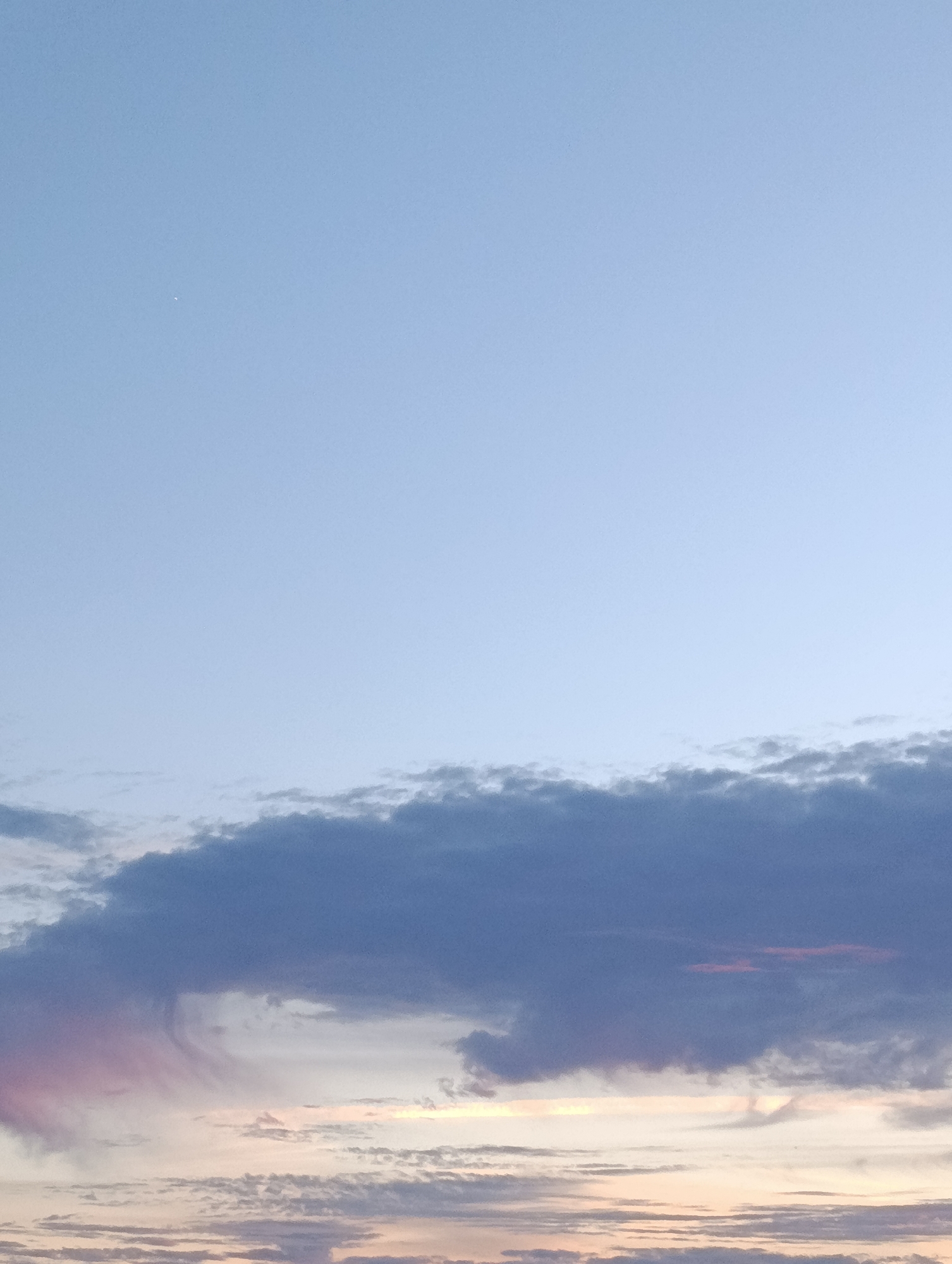 The image shows a dusky sky with scattered clouds over the city of Setúbal, Portugal. The sky is a gradient of pale blue at the top, transitioning to softer hues of pink and orange near the horizon, indicating either sunrise or sunset. A prominent band of dark, textured clouds stretches across the middle of the frame, obscuring the cityscape below. To the left, a wispy cloud formation exhibits a pinkish tint. The lower portion of the image reveals thinner layers of clouds with hints of color, suggesting the presence of scattered light. There are no discernible people, subjects, or objects in the frame other than the sky and clouds. The overall impression is one of natural atmospheric phenomena.