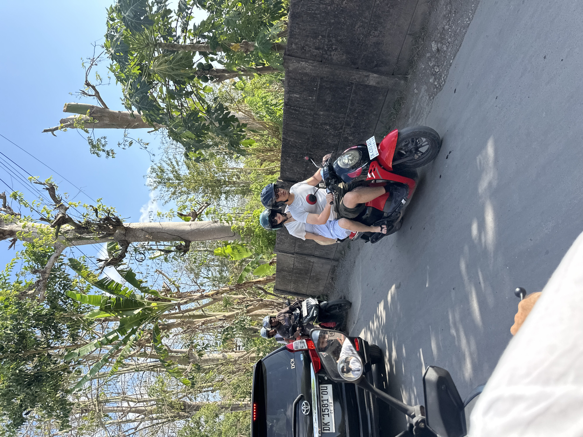 In Kuta Selatan, Indonesia, on a sunlit day, a man and a woman ride a red scooter through a street lined with trees and a concrete wall. The man, wearing a white t-shirt and dark shorts, drives the scooter, while the woman, in a white top and blue and white striped shorts, sits behind him. Both wear helmets. To their left, a black SUV is stopped in traffic, its license plate reading "DK 1581 OU". Another scooter with two passengers in helmets is visible behind the SUV. The road is paved, with shadows cast by the surrounding trees, and the sky is clear blue.