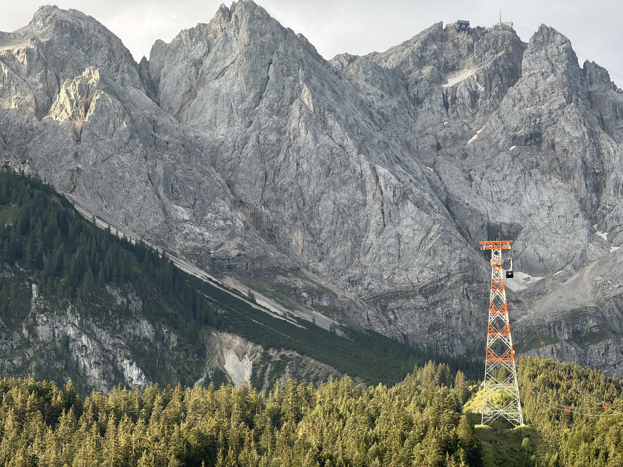 The image shows the Zugspitze, Germany's highest mountain, as seen from Grainau. In the foreground, a dense forest of pine trees covers a hillside. A tall, white and orange lattice tower, part of a cable car system, stands prominently in the mid-ground amidst the trees. Behind the tower, the rugged, grey slopes of the Zugspitze rise dramatically. The mountain is characterized by its steep, rocky faces, with some patches of snow visible near the summit. The sky is overcast. A cable car cabin is visible ascending towards the mountain peak, which is topped with a building complex and transmission towers. The scene conveys a sense of natural grandeur and human engineering in a mountainous environment.
