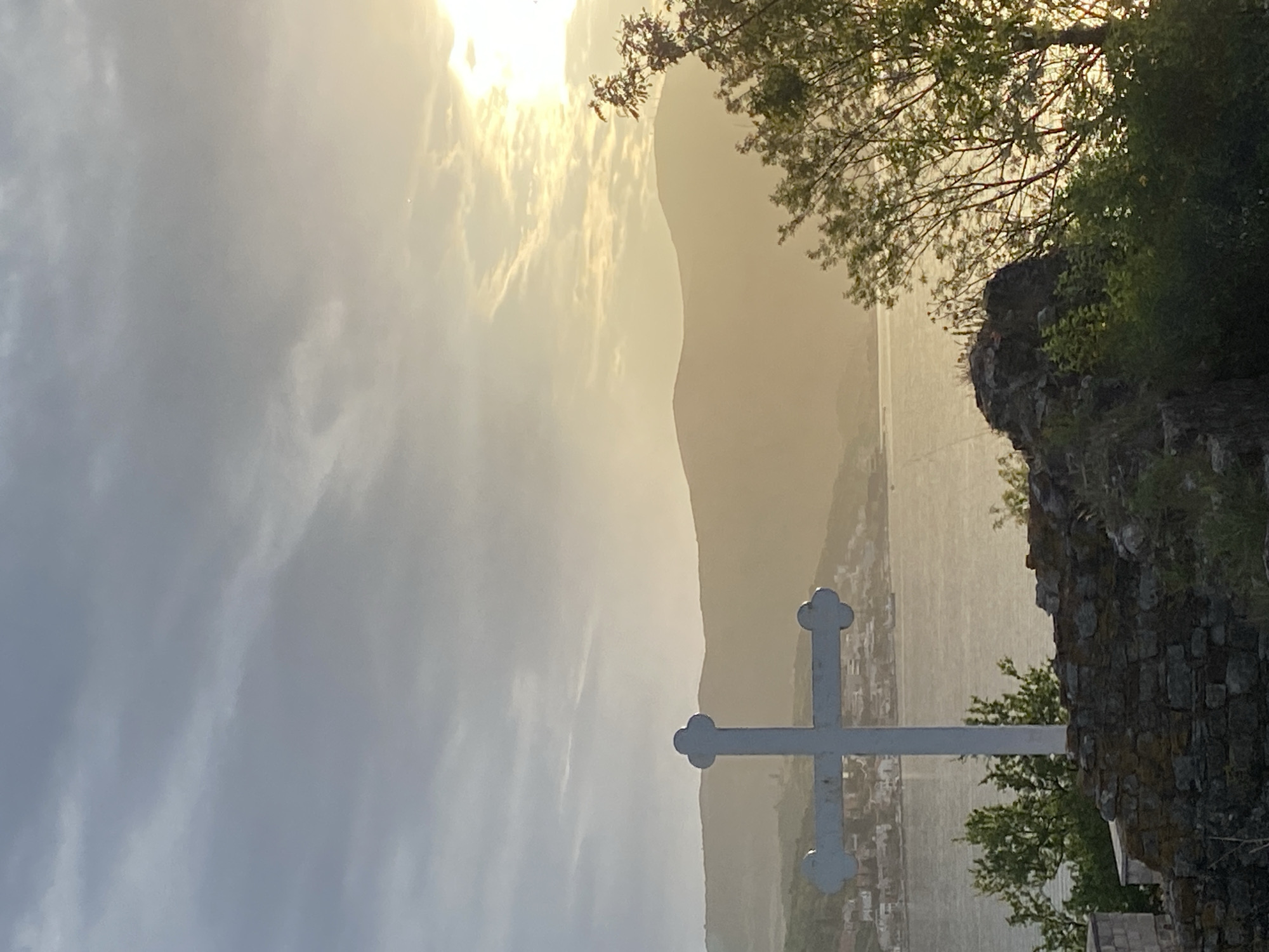 The image shows a landscape in Bogišići, Montenegro, dominated by a Christian cross in the foreground. The cross is white and appears to be made of metal. It stands near a stone structure or wall covered with vegetation. In the background, a body of water (likely the Bay of Kotor) is visible, with mountains rising on the opposite shore. The lighting is soft, possibly due to cloudy conditions or early morning light. The sky is bright and diffused, suggesting overcast weather. A town or settlement can be seen along the shoreline in the middle distance. A tree with green foliage is visible on the right side of the frame.