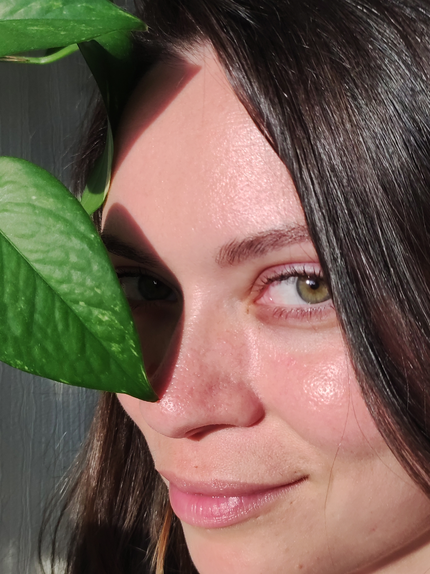 A close-up shot shows a young woman's face partially obscured by a green leaf. The woman has dark brown hair and fair skin with a rosy complexion. Her right eye is visible and appears to be hazel or green. The leaf is positioned on the left side of the frame, casting a shadow across the left side of her face. Her lips are slightly parted, revealing a hint of her teeth. The background is out of focus and appears to be a textured wall, possibly painted white. The lighting is bright and appears to be natural, highlighting the texture of her skin and the details of the leaf. No information about Chaikovsky, Russia is available in this image.