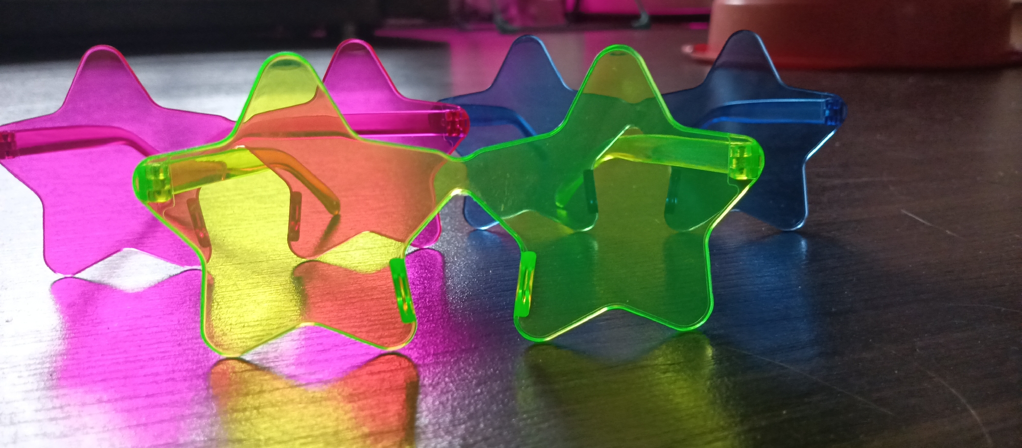 The image shows four pairs of star-shaped sunglasses arranged on a dark, reflective surface. The sunglasses are colored pink, yellow-green, and blue. The yellow-green glasses are in the foreground, partially obscuring the other pairs. The surface appears to be a table or desk. The location is unknown.