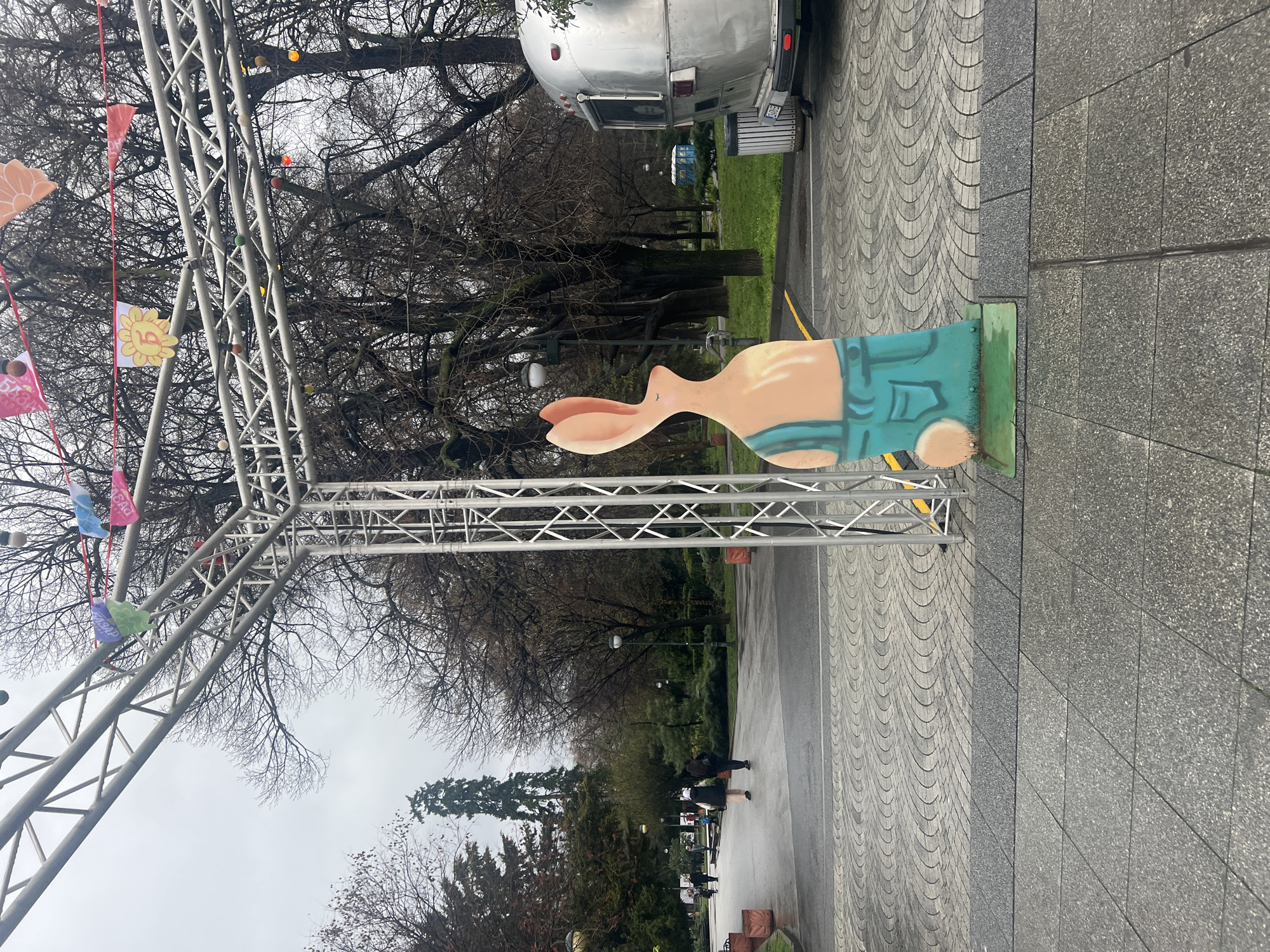 The scene depicts a public space in Plovdiv, Bulgaria, possibly a park or pedestrian area. A silver Airstream trailer is parked adjacent to an event setup, characterized by a metal truss structure adorned with colorful pennants or flags. A cartoonish, cutout figure of a rabbit wearing overalls is positioned prominently. The ground is paved with a patterned design, transitioning to a plain gray surface. Trees with bare branches fill the background, and figures of people can be seen walking in the distance. The day appears overcast.