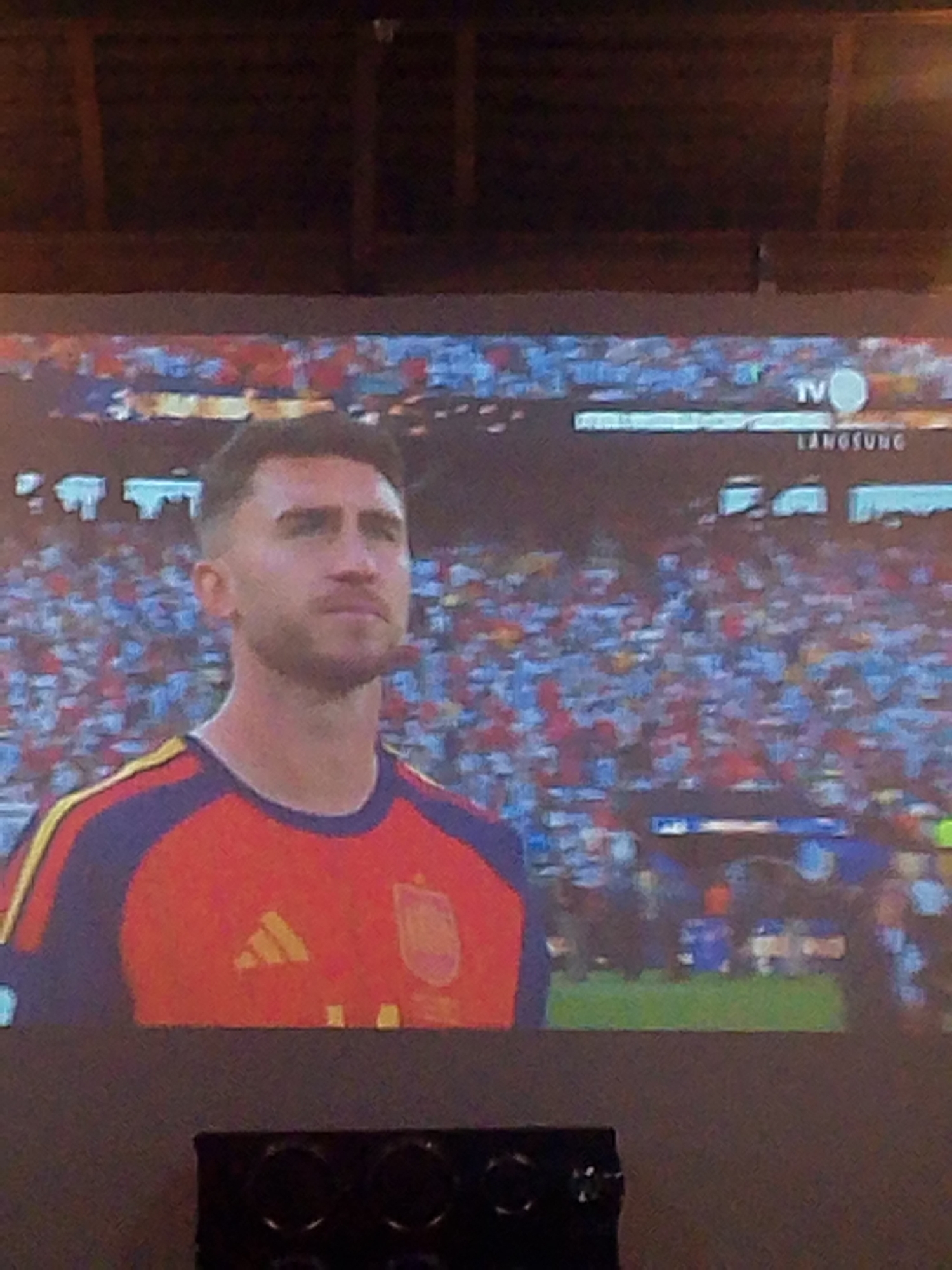 A man, who appears to be a soccer player wearing a red and yellow jersey, is the primary subject of the image. He is looking to the right with a neutral expression. Behind him, a large crowd fills a stadium, with many people wearing red and blue clothing. The scene is likely a soccer match. In the top right corner of the image, a TV logo and the word "LANGSUNG" (which means "live" in Indonesian) are visible, suggesting the image is a broadcast. The setting appears to be a stadium in Meunasahmee, Indonesia.