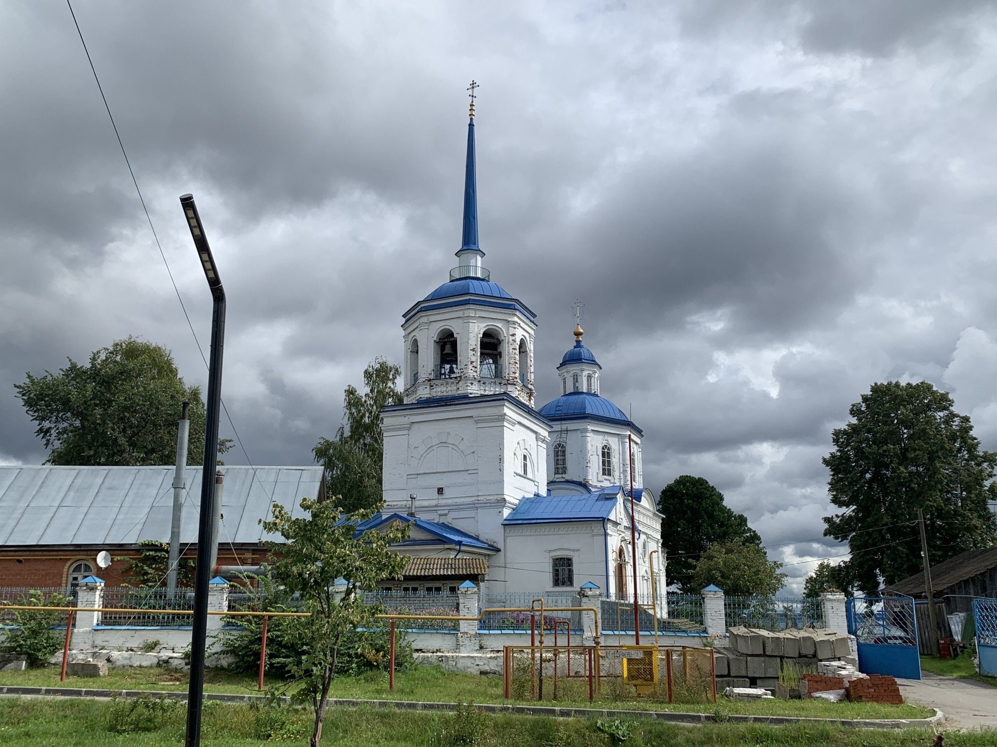 The image shows a white church with blue domes and a tall, slender blue spire topped with a cross. The church has a bell tower attached to its front. The sky is overcast with heavy, dark clouds. In the foreground, there is a black lamppost and a section of a building with a gray metal roof. A low fence, partially made of blue and white brick pillars with yellow and red horizontal bars, surrounds the church property. There are trees on either side of the church, and a small amount of construction material, including bricks and concrete blocks, is piled near the fence on the right side of the image. The environment appears to be a rural or suburban area in the city of Malaya Romanova, Russia.
