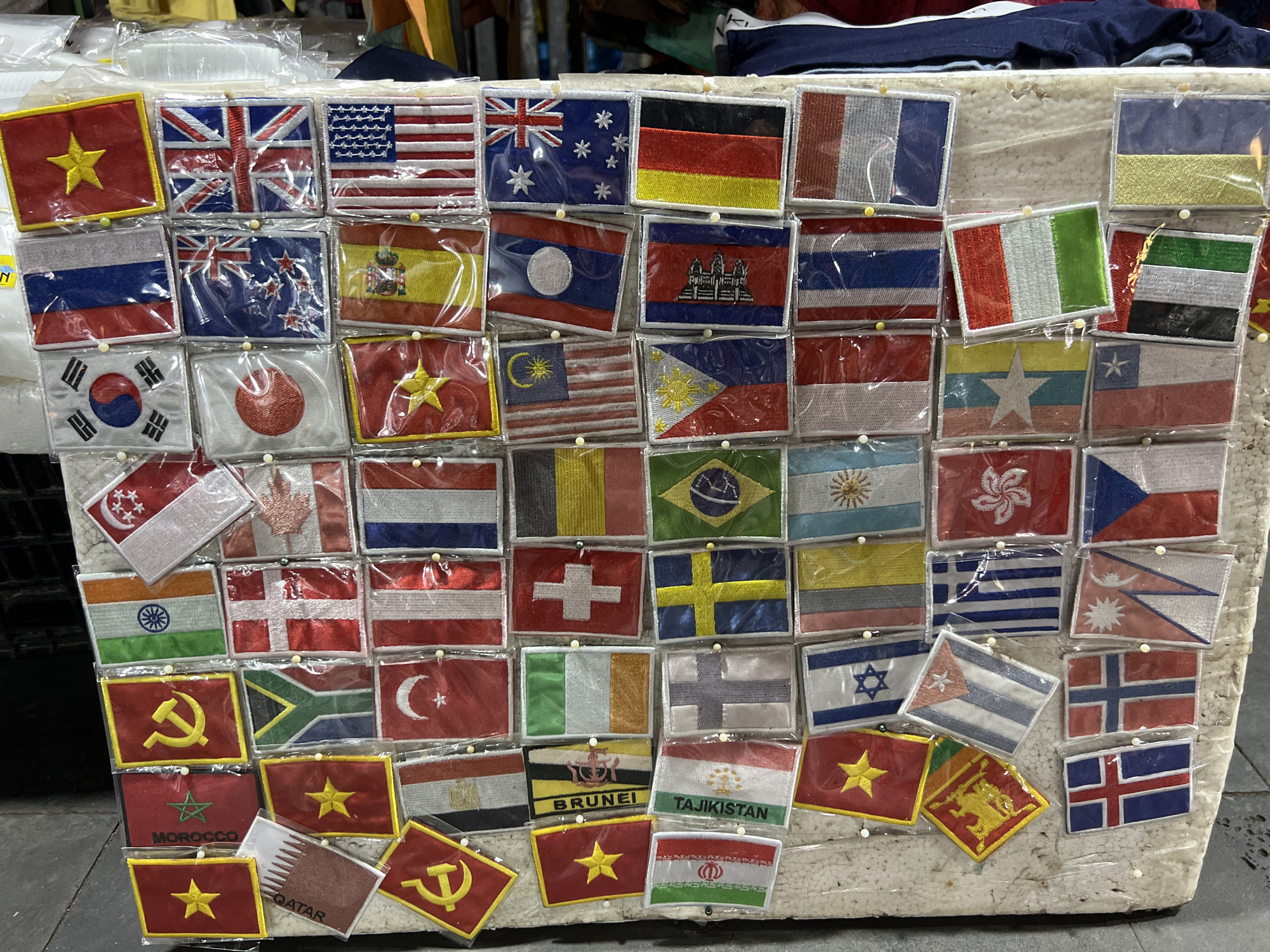 The image displays a densely packed arrangement of small fabric flag patches, predominantly featuring national flags. These patches are individually packaged in clear plastic and pinned to a textured, off-white surface, possibly a display board or styrofoam. The flags are varied in design, with recognizable patterns from countries such as Vietnam, the United Kingdom, the United States, Russia, Australia, Germany, and many others. Some patches have text labels, like "MOROCCO" and "QATAR." The background suggests a retail environment with glimpses of clothing and other merchandise. The overall impression is of a stall or market display, likely in a tourist area, given the variety of international flags. The scene is static, with no visible people or active interactions. The lighting is even, allowing for clear visibility of the details on each flag patch. The environment appears to be indoors or covered, with a focus on the product display.