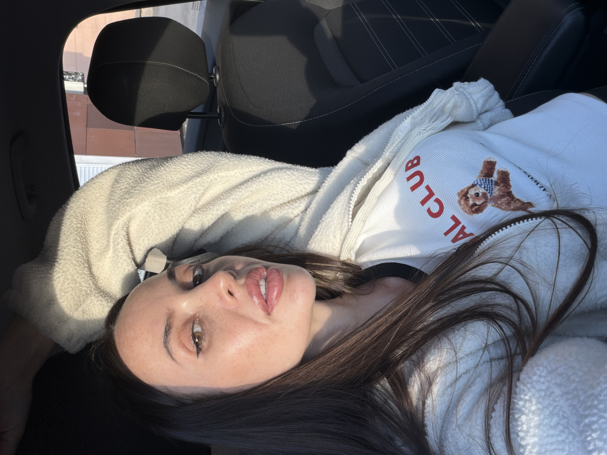 A woman with long, dark hair and full lips is inside a car. She is wearing a white fuzzy jacket over a white t-shirt that has red lettering saying "AL CLUB" and a picture of a dog wearing a bandana. The sun is shining, casting shadows on her face and in the car. The background shows a car headrest and a building with brown siding.