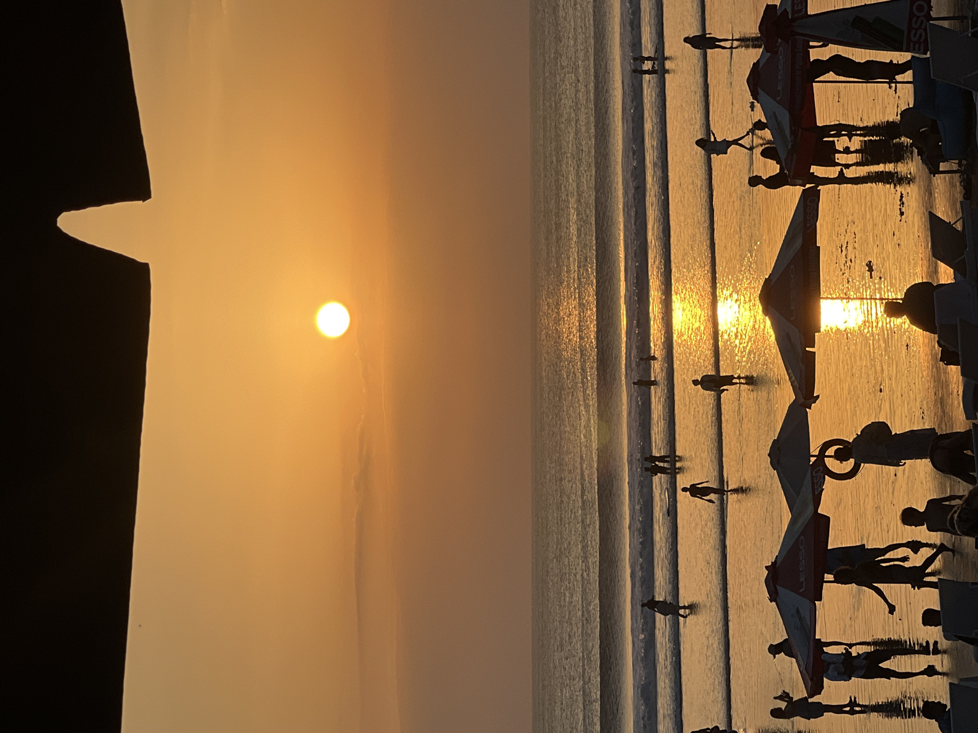 A sunset scene at Kuta Beach, Bali, Indonesia, features the sun low on the horizon, casting a warm orange glow across the sky and reflecting on the wet sand. Silhouettes of beachgoers are visible, with some wading in the gentle waves and others walking along the shore. Several beach umbrellas and lounge chairs are scattered on the sand in the foreground. The image captures the relaxed atmosphere of a beach at dusk.