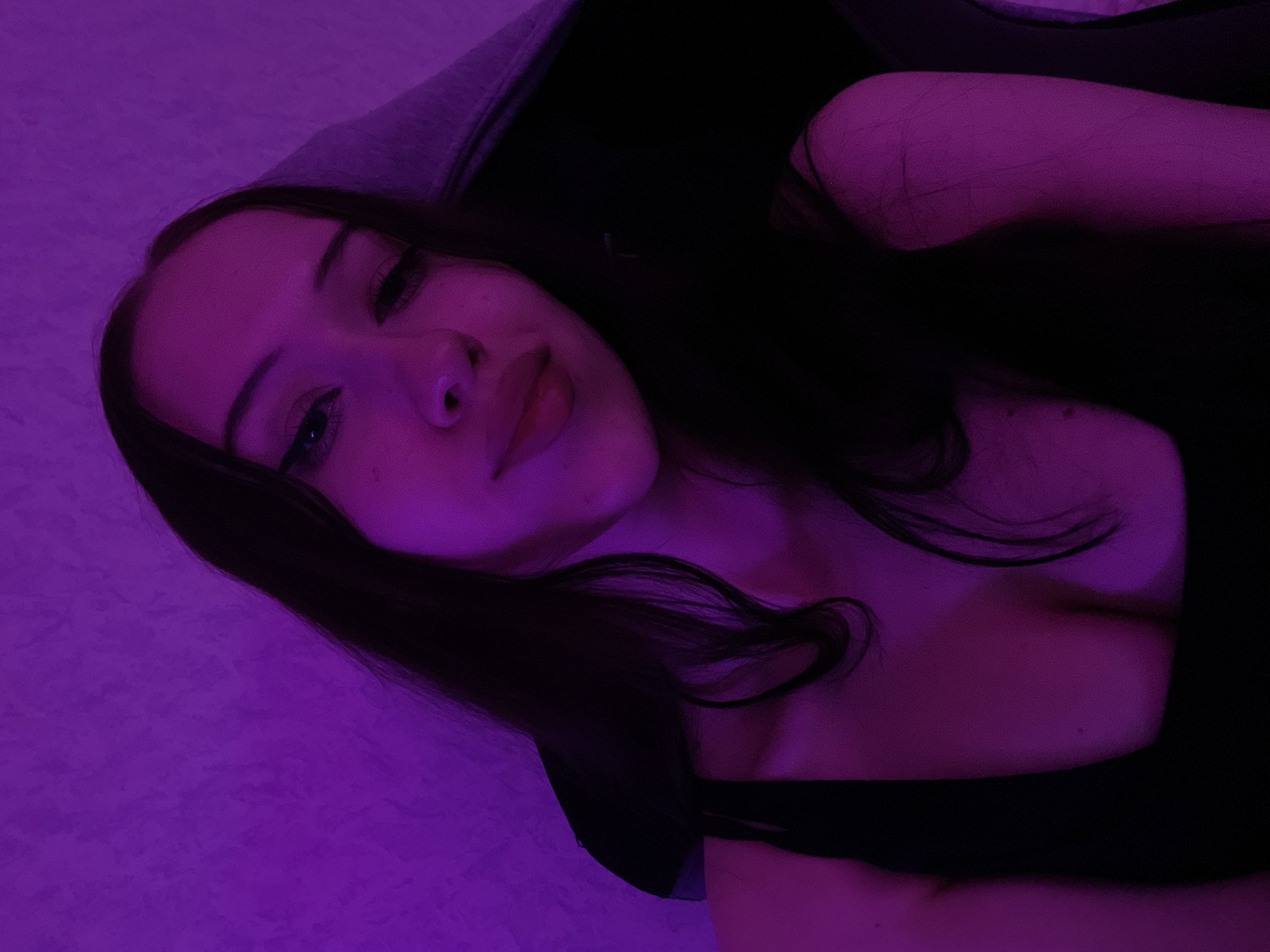 The image shows a young woman indoors, lit by purple light. She has long, dark hair and is wearing a black top. The background appears to be a textured wall. The location in Votkinsk, Russia, is not discernible from the image.
