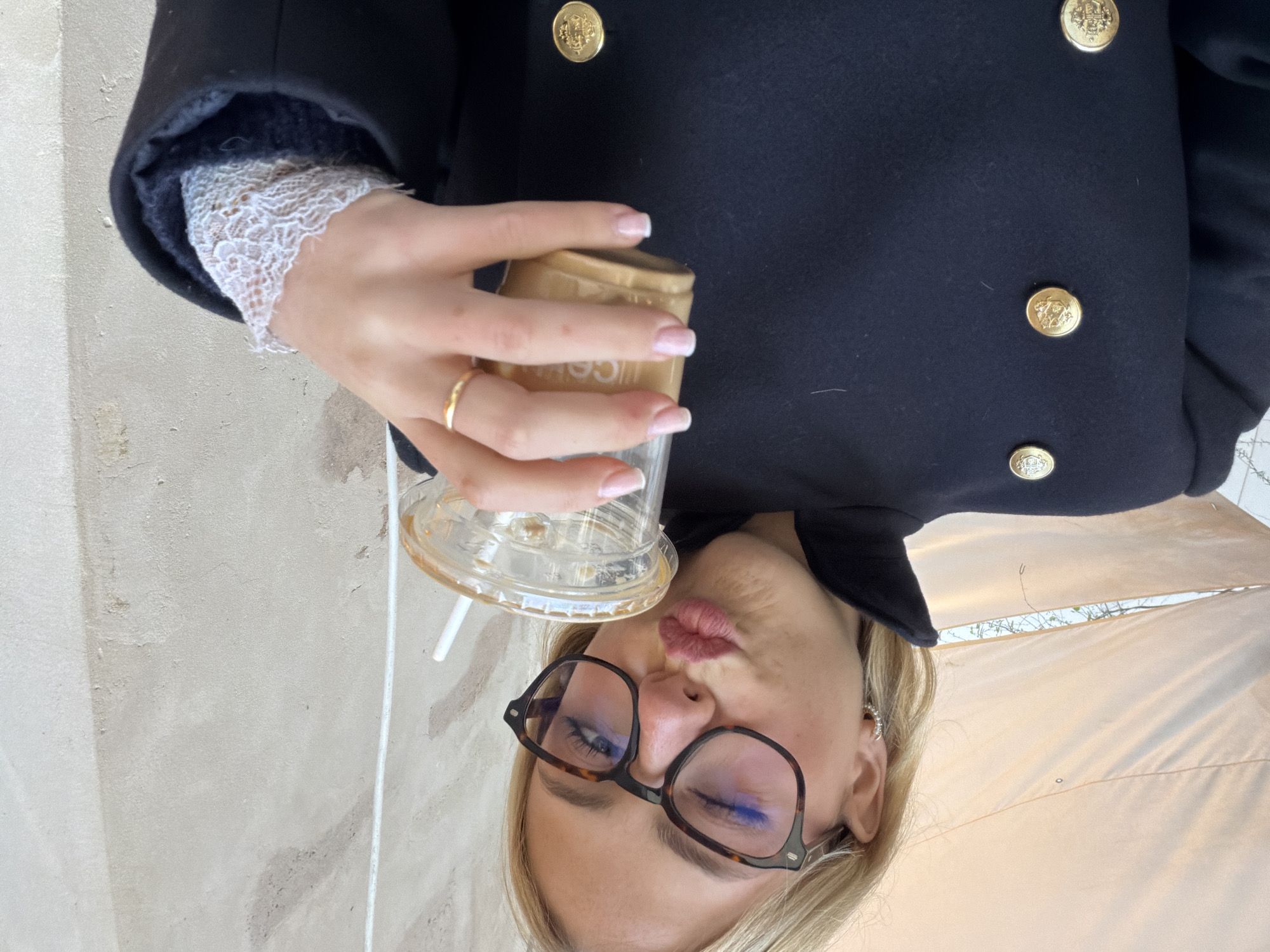 A blonde female is depicted in a close-up, selfie-style shot, winking with her left eye and puckering her lips. She wears brown tortoise-shell eyeglasses and a dark, likely navy or black, double-breasted coat adorned with gold-toned buttons featuring embossed crests. A white lace cuff is visible on her right wrist. She holds a clear plastic cup containing an iced, light brown beverage, presumably coffee, with a white straw inserted. A plain gold band ring is visible on the ring finger of her right hand, and her fingernails exhibit a French manicure. The immediate background on the left consists of a light beige or off-white fabric canopy or awning. To the right and behind the subject, a textured, light-colored stone or stucco wall, indicative of older European architecture, completes the scene. The architectural and atmospheric elements are consistent with an outdoor cafe or street setting in Paris, France.