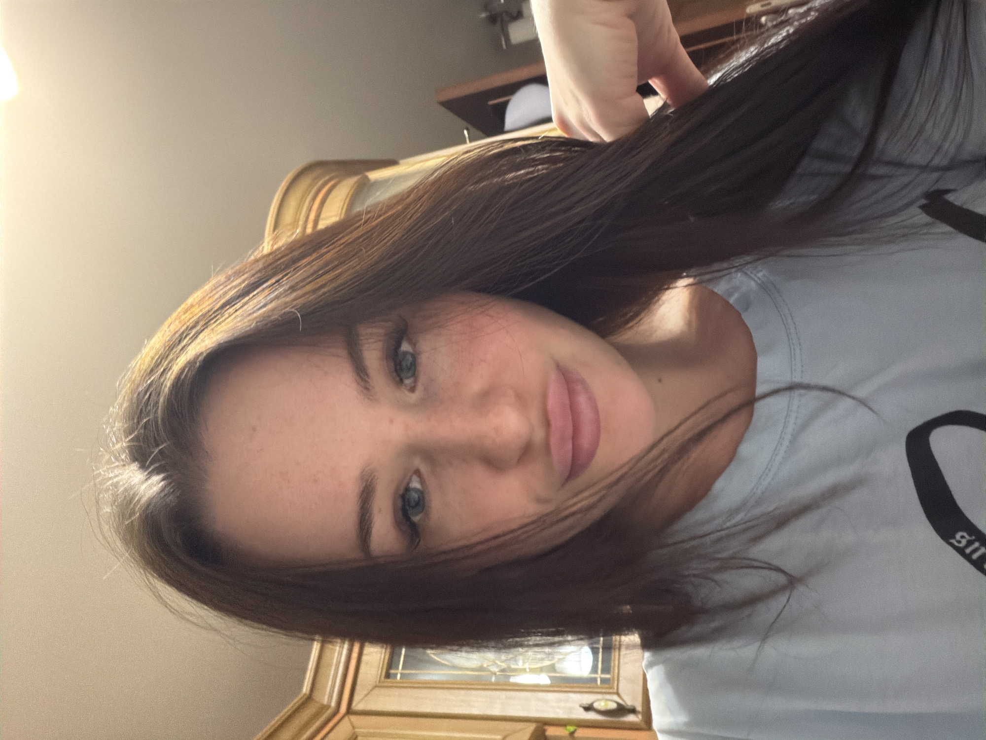 The image features a young female taking a selfie indoors. She is positioned horizontally in the frame, looking directly at the camera with a neutral to slightly smiling expression. Her left hand is visible at the top, gently holding and lifting a section of her long, dark brown hair. Her hair appears to have some lighter brown or auburn highlights, particularly noticeable around her face.
She has fair skin with visible freckles across her nose and cheeks. Her eyes are blue, and her lips appear full, possibly enhanced with a light lip gloss or lipstick. She is wearing a light blue or gray t-shirt. On the right side of her shirt, partially visible text in a dark, stylized font can be seen, which appears to spell "girls" or a similar word.
The background suggests an indoor setting, likely a home. To the left, a bright light source is visible, possibly from a lamp. Behind her, there is wooden furniture, resembling a cabinet or dresser, with a reflective surface that might be a framed picture or mirror. A white object, possibly a cap, is visible on a shelf in the background. The overall lighting is warm.
The image captures an intimate, casual moment, likely a self-portrait taken in a personal space.
Location context: Unknown, Russia.