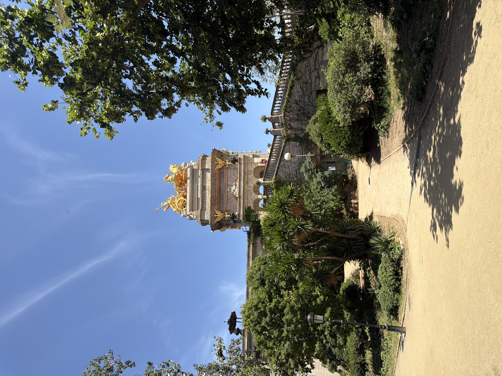 The image shows a view of the Monumental Cascade at Parc de la Ciutadella in Barcelona, Spain. The monument, made of stone and adorned with golden sculptures, is visible in the background. A chariot pulled by horses is perched atop the monument, with a winged figure standing in the chariot. Green trees and foliage surround the monument, and a sandy path leads through the park. The sky is clear and blue with wispy clouds. The scene is bathed in sunlight, casting shadows on the path.