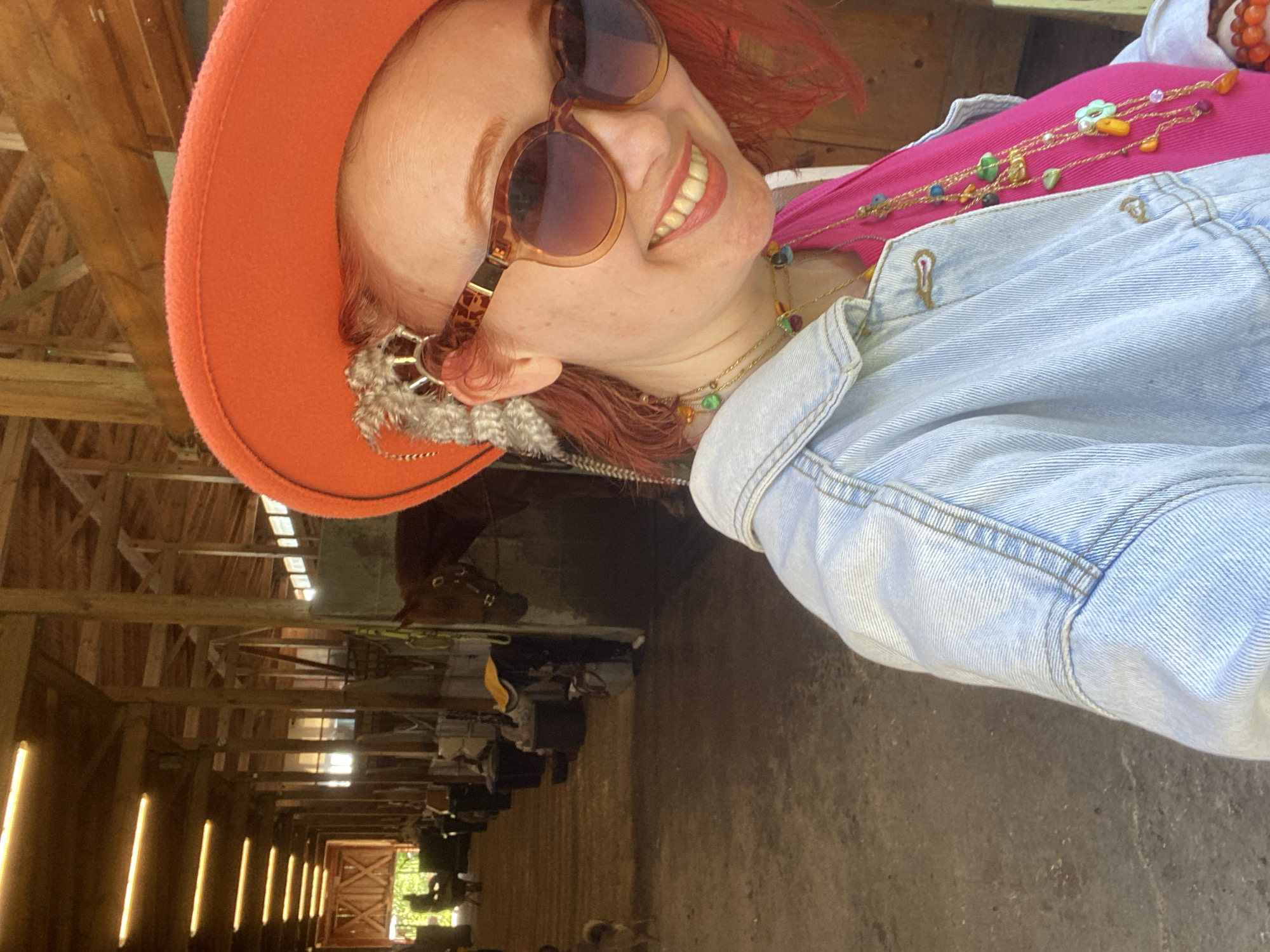 A woman wearing a burnt orange hat and sunglasses is smiling at the camera. She is wearing a pink shirt and a light denim jacket. Several gold necklaces with colorful beads are visible. The woman appears to be standing in a stable. There are horses in stalls visible in the background. The stable has a wooden frame and roof. The ground is dirt or concrete. Light enters the stable through vertical openings in the wall. The location is unclear, but no evidence suggests Прогресс, Russia.
