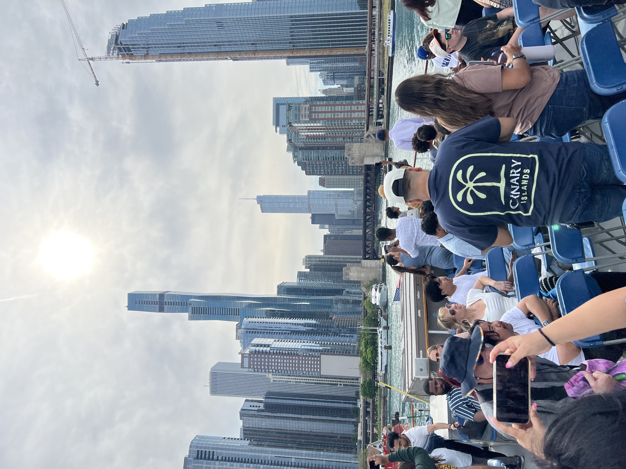The image depicts a view of the Chicago skyline from a boat on the Chicago River. The sky is overcast, with the sun shining through the clouds. Numerous skyscrapers dominate the background. A construction crane is visible atop one of the taller buildings. In the foreground, there is a group of people sitting in blue plastic chairs on what appears to be a tour boat. Some people are taking pictures with their phones, while others are looking at the buildings. A man is wearing a navy blue shirt with a 'Canary Islands' graphic. The overall scene captures a typical tourist experience of taking a river tour to view the architecture of Chicago.