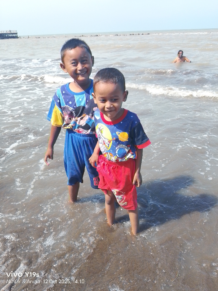 The image depicts a sunlit coastal scene in Limbangan, Indonesia. In the foreground, two male children stand in shallow, murky brown water. The taller child, positioned to the left, wears a blue and yellow t-shirt with cartoon characters and blue shorts, smiling directly at the viewer. The shorter child, positioned to the right, wears a blue t-shirt with a space-themed design and red shorts, looking slightly downward and to his right. Both children are barefoot, with water reaching their lower legs, and their shadows are visible behind them in the water. Further out in the water, an adult male, bare-chested, is partially submerged, visible from the chest up, looking towards the left. The background features a calm, grayish-brown sea under a clear blue sky. A dark, pier-like structure is visible on the far left of the horizon, and a line of dark breakwater elements extends horizontally across the water in the mid-distance.